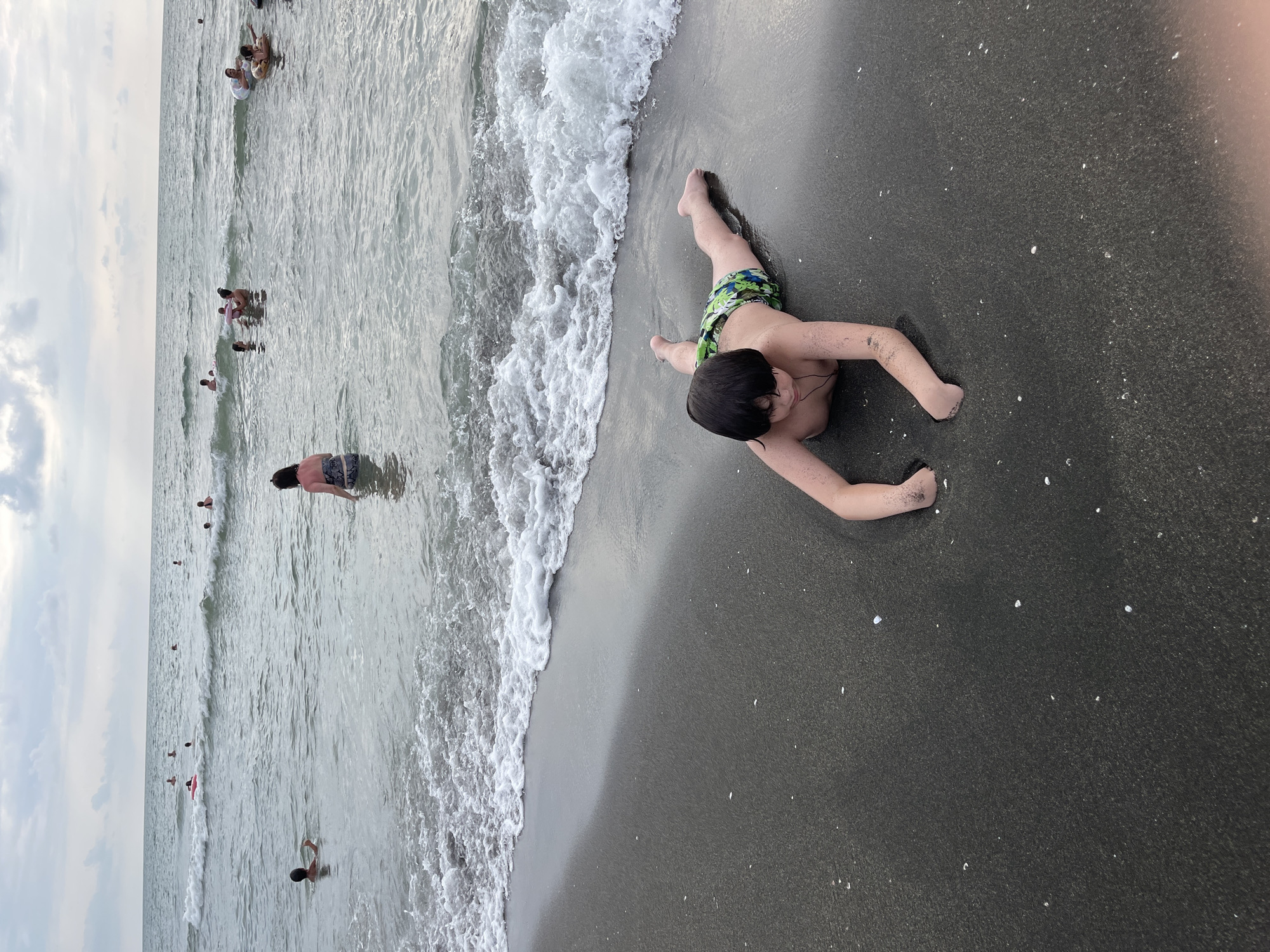 A young boy in green and blue swim trunks lies face down on the dark sand at the edge of the Black Sea in Ureki, Georgia. His arms are extended forward, and his hands are partially submerged in the wet sand. The shallow water of the sea laps at the shore, creating white foam. In the background, numerous people are visible swimming and wading in the water. The sky is overcast with clouds.