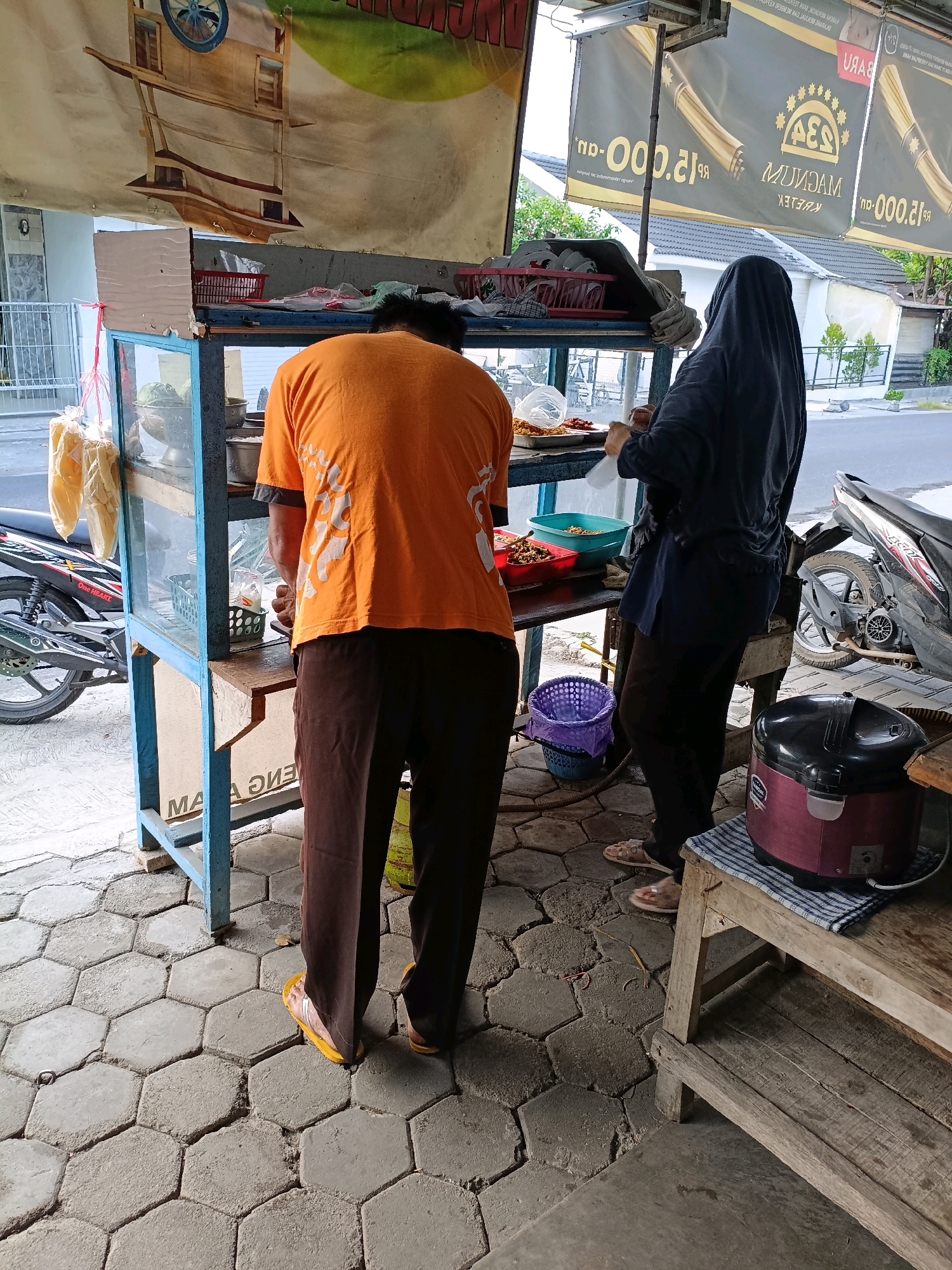 Two individuals are observed near a food stall, situated on a hexagonal paver sidewalk in Maguwoharjo, Indonesia. The stall features a glass display case filled with various food items, including what appears to be fried items in red trays and possibly vegetables in a metal bowl. A person wearing an orange t-shirt and brown pants stands facing the stall, seemingly engaged in preparing food. To the right, a person in a dark headscarf and navy blue clothing is seen holding a plastic bag and interacting with the food items within the stall. A purple laundry basket with a plastic bag inside is placed on the ground to the left of the stall. In the background, a motorcycle is parked to the left of the stall. A rice cooker and a wooden shelf are positioned to the right of the stall. Above the stall, banners advertising a product with the text "MAGNUM KRETEK" and prices are displayed. The overall scene depicts a bustling street food vendor environment.