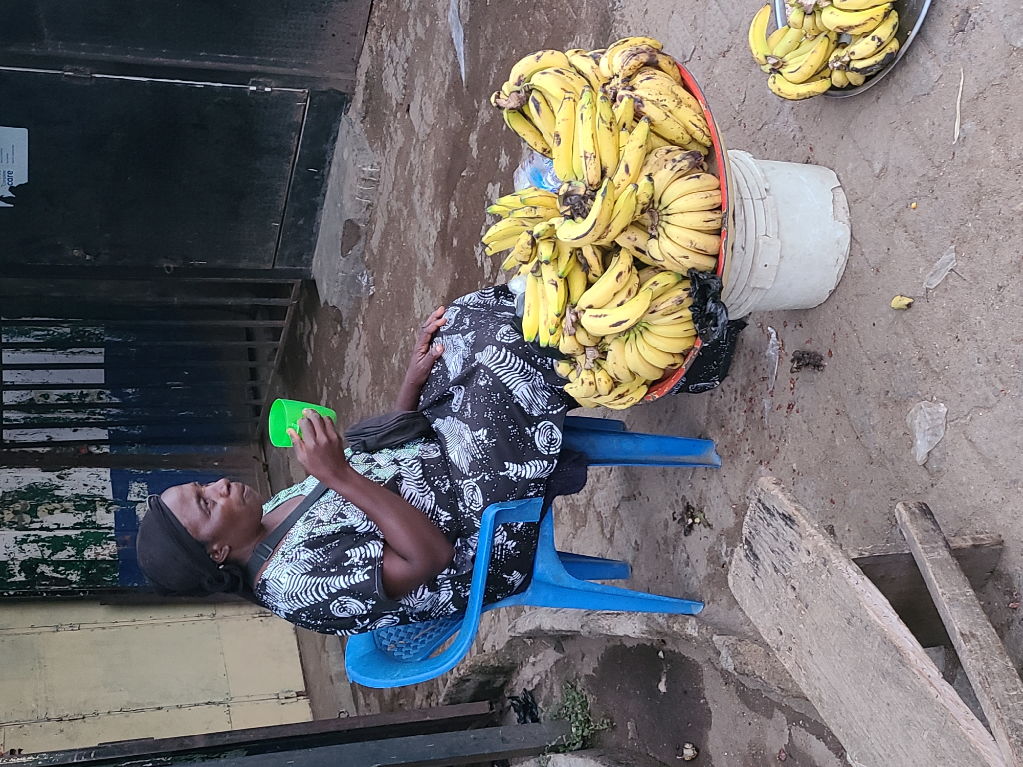 A Black woman, wearing a black headscarf and a black and white patterned dress, sits on a blue plastic chair. She holds a green plastic cup in her right hand, bringing it to her mouth as if drinking. The woman is positioned to the left of the frame. In front of her, to the right, is a large platter filled with ripe yellow bananas. This platter is resting on a white bucket. To the right of the platter, a second, smaller platter also contains a pile of bananas. The ground is dusty and uneven, with scattered debris. In the background, a closed metal gate with vertical bars is visible, set against a weathered wall. A portion of a sign with the word "care" is visible on the wall above the gate. The scene appears to be an outdoor market or street vendor stall in Jos, Nigeria.