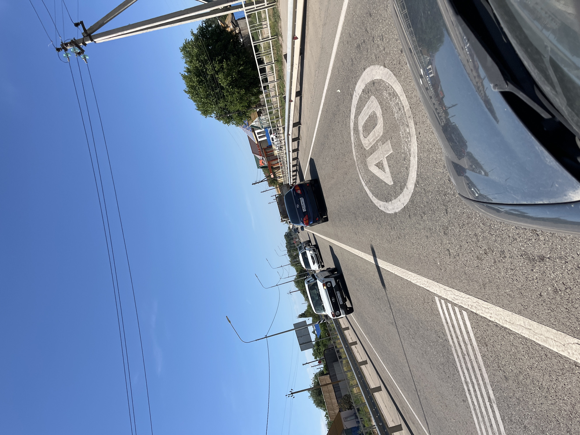 The image depicts a view from inside a vehicle traveling on a two-lane road in Bakhtemir, Russia, under a clear blue sky. The road is bordered by a metal guardrail and a sidewalk with houses and trees visible on the right side. A speed limit sign of 40 is painted on the road surface. Several vehicles are on the road, including a white Lada Niva in the foreground, followed by other cars. Power lines and poles are visible above the road and along the roadside, suggesting it is a populated area. The sun is bright, casting shadows. The overall impression is a typical scene of road travel in a semi-urban or rural environment.