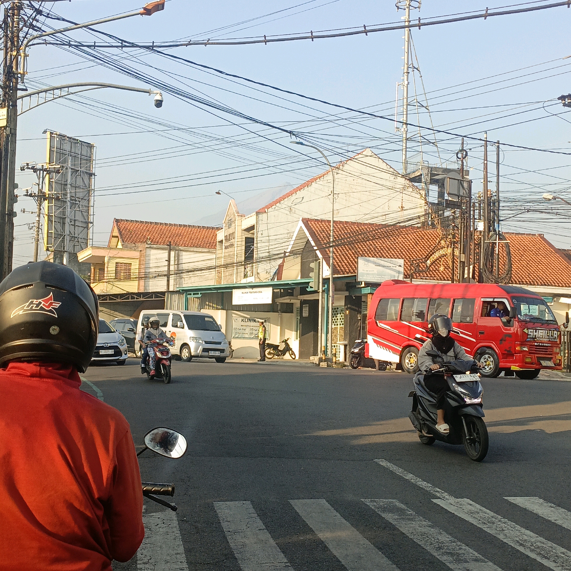 This image captures a street scene in Salatiga, Indonesia, likely during daytime given the clear sky and warm lighting. The foreground is dominated by the back of a person wearing a red jacket and a black helmet, who is on a motorcycle, facing away from the camera. Their motorcycle's side mirror is visible to the right.
In the middle ground, several vehicles are present on the asphalt road. A white van is parked on the left, and another white van is moving on the right. A red minibus with "SHEILA" written on its side is also present. Several motorcycles are also visible, including one with a rider in a grey jacket and black pants, wearing a helmet and a black face covering, stopped at a crosswalk. Another motorcycle is seen with a rider wearing a patterned outfit and a helmet.
The background features buildings with orange tile roofs and beige walls. Utility poles with numerous overhead wires are prominent throughout the scene, indicating a developed urban area. A traffic camera is mounted on an arched pole above the road. Signs are visible on some of the buildings, one appearing to be a clinic entrance with text that includes "KLINIK PRATAMA DEWI". The overall atmosphere suggests a busy intersection or a commercial street.