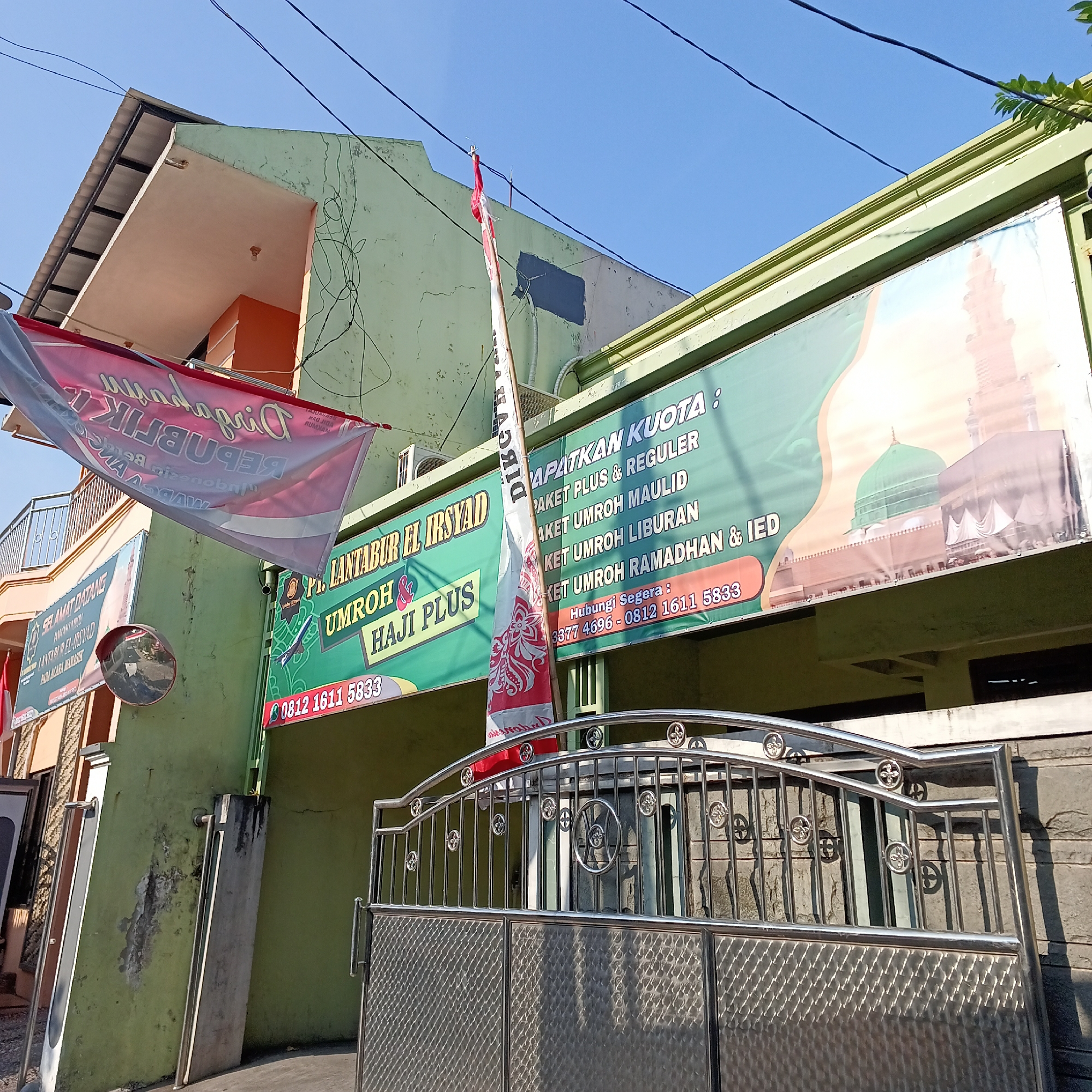 The image depicts the exterior of a building in Wedoro, Indonesia, likely an office or travel agency specializing in Umrah and Hajj pilgrimages. A large green banner displays text in Indonesian related to Umrah packages, featuring images of Islamic holy sites. To the left, a red and white banner hangs, partially obscuring another sign that reads "PT. LANTABUR EL IRSYAD UMROH & HAJI PLUS". A Polish flag is prominently displayed on a pole in front of the building. A metal gate with intricate designs is in the foreground, with a reflective convex mirror mounted on the wall to the left. The sky is clear blue, suggesting a sunny day. The building itself appears to be a light green color, with some signs of wear and tear. Electrical wires are visible crisscrossing the facade.