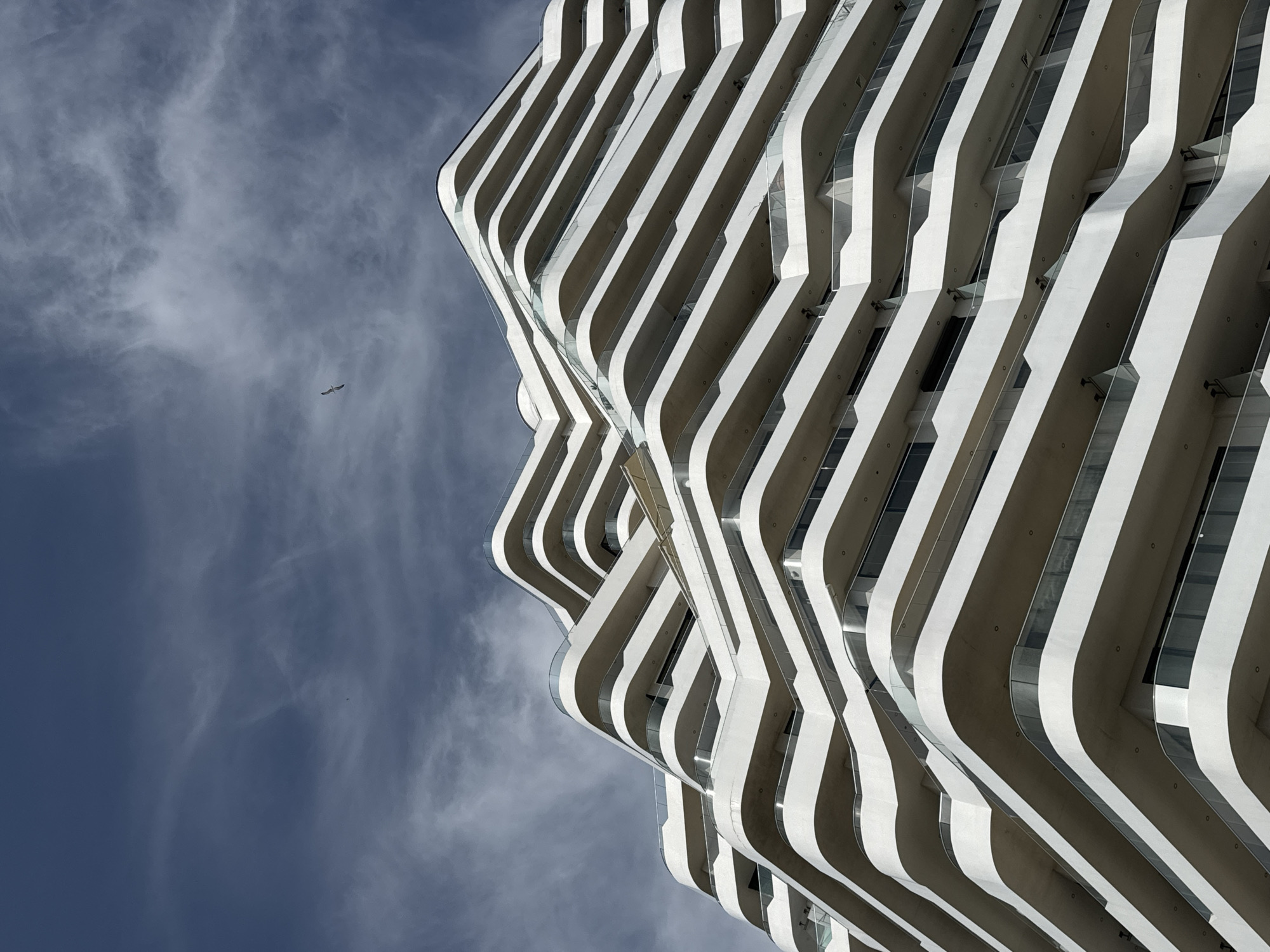 The photograph captures a portion of a modern building with a unique facade design, characterized by repeating white and grey stripes forming curved balconies. The sky is visible on the left, with faint streaks of clouds. A small bird is flying across the sky. The building is situated in Burgas, Bulgaria.