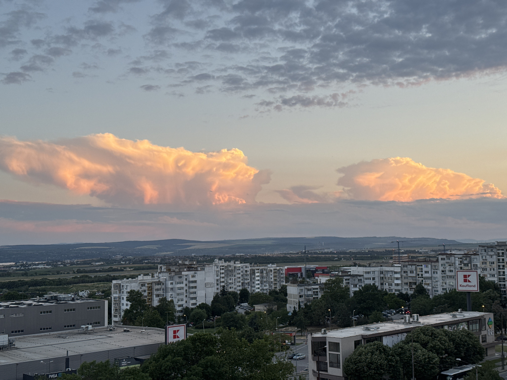 The image depicts a cityscape in Varna, Bulgaria, during what appears to be sunset or sunrise, characterized by a sky filled with dramatic, illuminated clouds. The predominant colors in the sky range from soft blues and grays to vibrant oranges and pinks, suggesting a significant atmospheric event.
In the foreground and mid-ground, a dense urban environment is visible, comprised of apartment buildings and commercial structures. These buildings are predominantly rendered in shades of white and gray, with visible balconies and window arrangements characteristic of Eastern European architecture. Lush green trees are interspersed throughout the urban landscape, softening the architectural rigidity.
Notable features include a prominent red and white sign with the "Kaufland" logo, indicating a commercial presence. Several rooftops are visible, some featuring industrial equipment like air conditioning units. A large, gray industrial building with numerous windows dominates the left side of the frame. In the distance, a sprawling landscape of rolling hills and vegetation stretches towards the horizon, dotted with more structures and hints of water, possibly a bay or river. Construction cranes are also visible in the far distance, suggesting ongoing development.
The overall impression is of a city at the edge of evening or dawn, with natural atmospheric beauty contrasting with the man-made urban environment. No people are distinctly visible in the image.