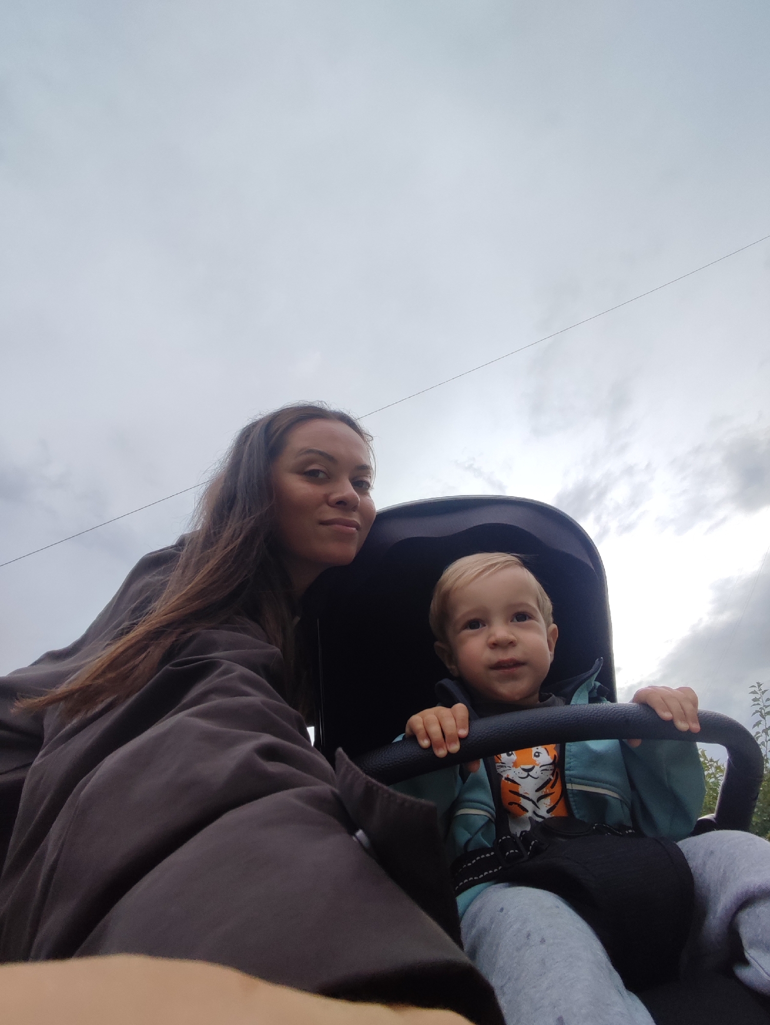 The image shows a woman and a young child in a stroller, taken outdoors under a cloudy sky. The woman is on the left, wearing a dark, oversized jacket and looking towards the camera with a slight smile. Her long, brown hair is loose. The child is in the stroller on the right, wearing a light blue jacket over a t-shirt with a tiger graphic, and grey sweatpants. The child is looking forward with a curious expression, holding onto the stroller's handlebar. The background consists of a gray sky with some visible clouds and a thin wire stretching across it. The scene appears to be a casual outing, possibly in a park or a residential area, but no specific landmarks of Chaikovsky, Russia are visible.