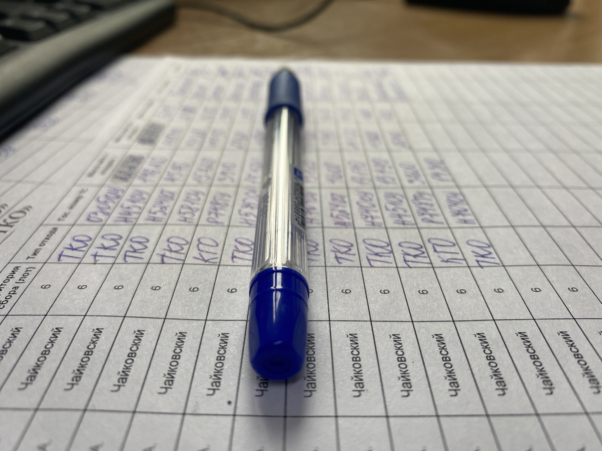 A ballpoint pen is centered on top of a piece of paper with writing on it. The writing on the paper appears to be in Cyrillic script, specifically in Russian. The name "Чайковский" (Chaykovskiy) is consistently written in the first line. The first column appears to be a description, the second is a number (6) and the third is an acronym (TKO). The paper is most likely a form. The photograph is taken at an eye-level point of view. Due to the writing on the paper mentioning "Чайковский" (Chaykovskiy) and the Russian script, it can be inferred that this image may be taken in a location related to or associated with the city or a place in Ol'khovskoye, Russia, where people may be filling out administrative or reporting forms.