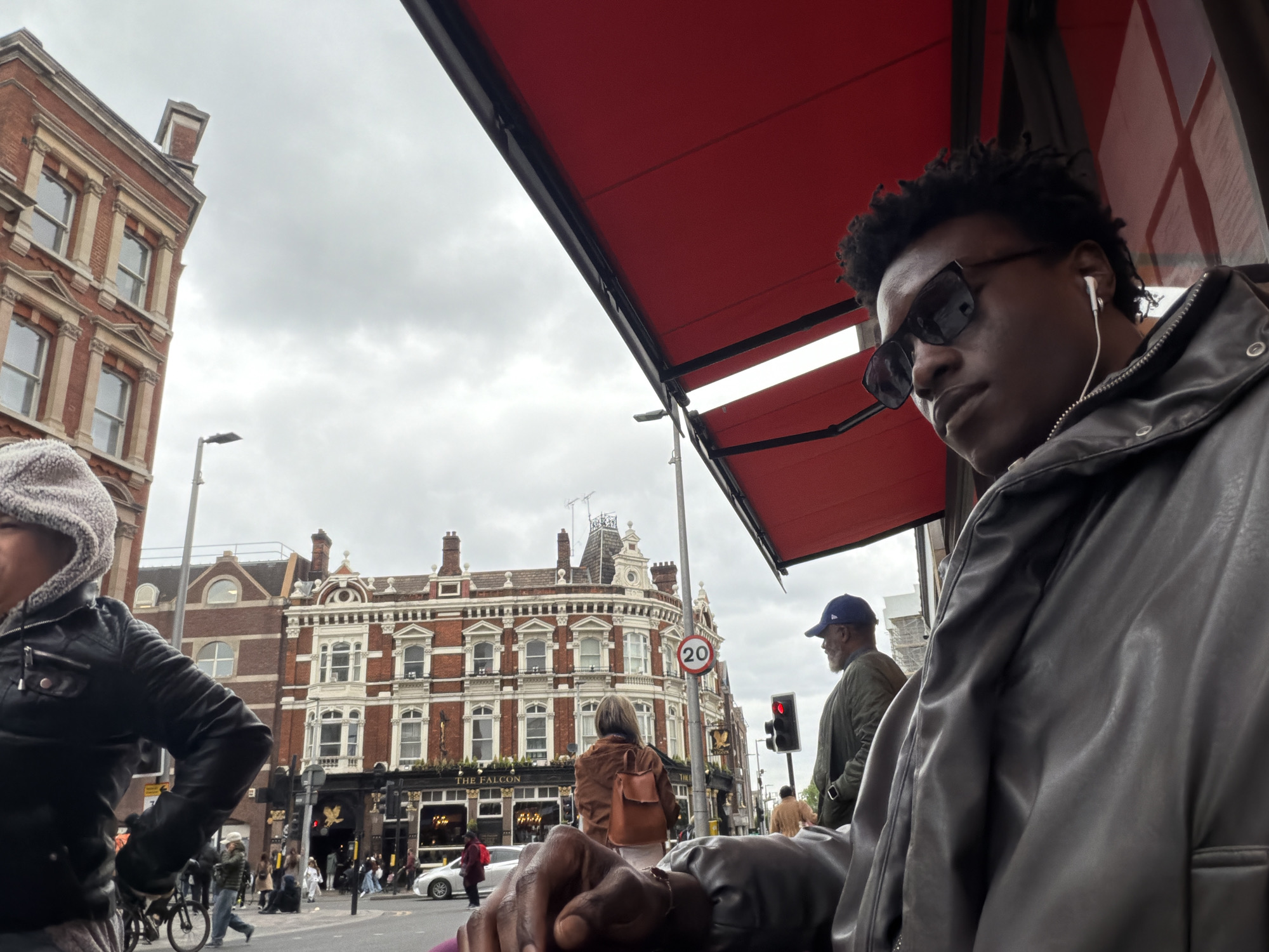 The image shows a street scene in London, United Kingdom. The foreground is dominated by two individuals, one a young man with sunglasses and earbuds, wearing a leather jacket. The other individual is to the left, partially obscured. The background reveals a red-brick building with white detailing. "THE FALCON" can be seen written on the storefront. There are pedestrians crossing a street in front of the building. The sky is overcast. The photograph appears to have been taken from under a red awning. There is a 20 speed limit sign present. A traffic light is visible with a red light illuminated.