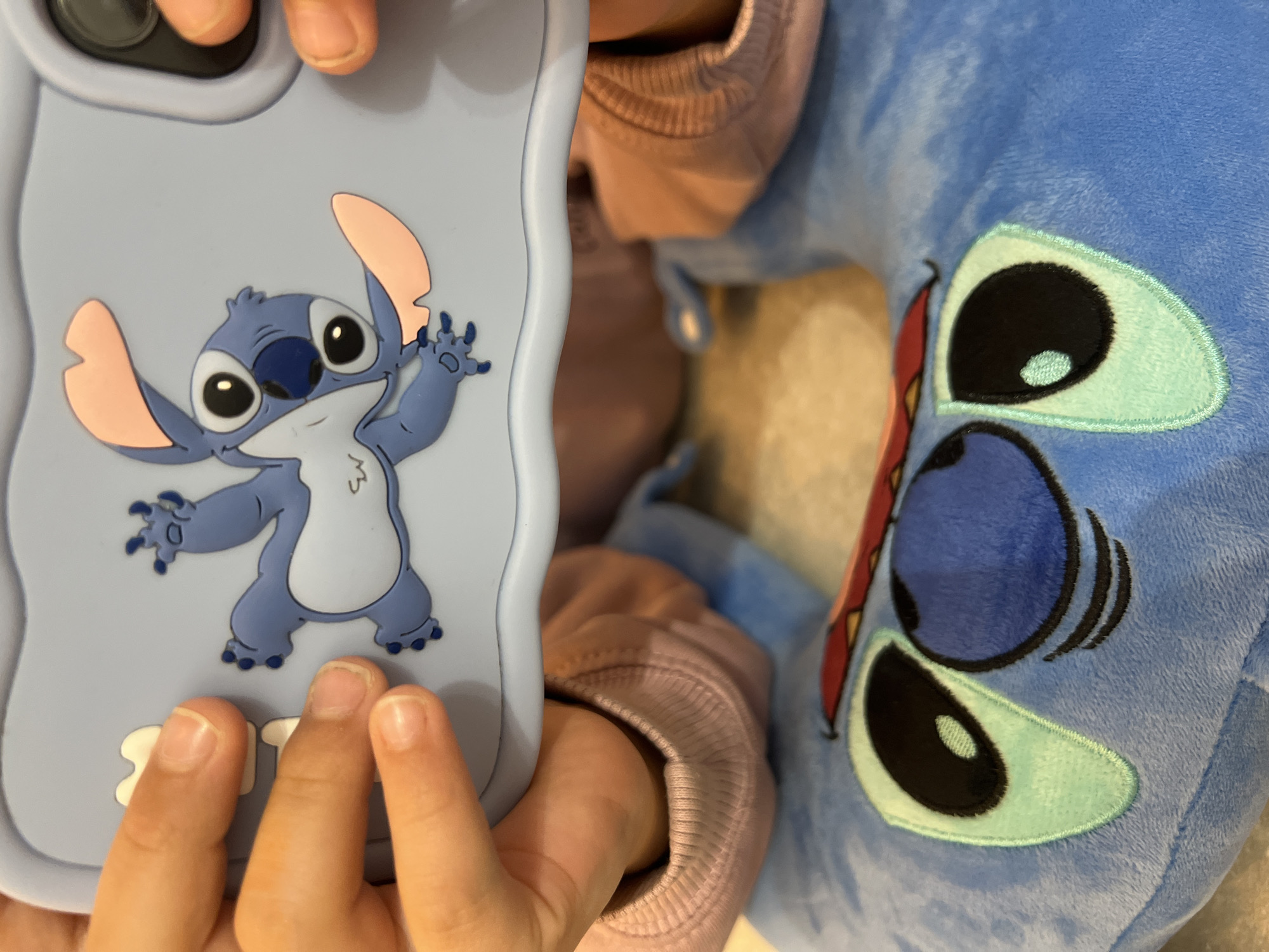A close-up captures a child holding a light blue phone case with a cartoon rendition of Stitch on it. The phone case is held in front of a blue, plush Stitch toy. The child's hands and a sleeve of a pink sweatshirt are also visible. The location and city of Budva, Montenegro are not discernible from the visual information provided.