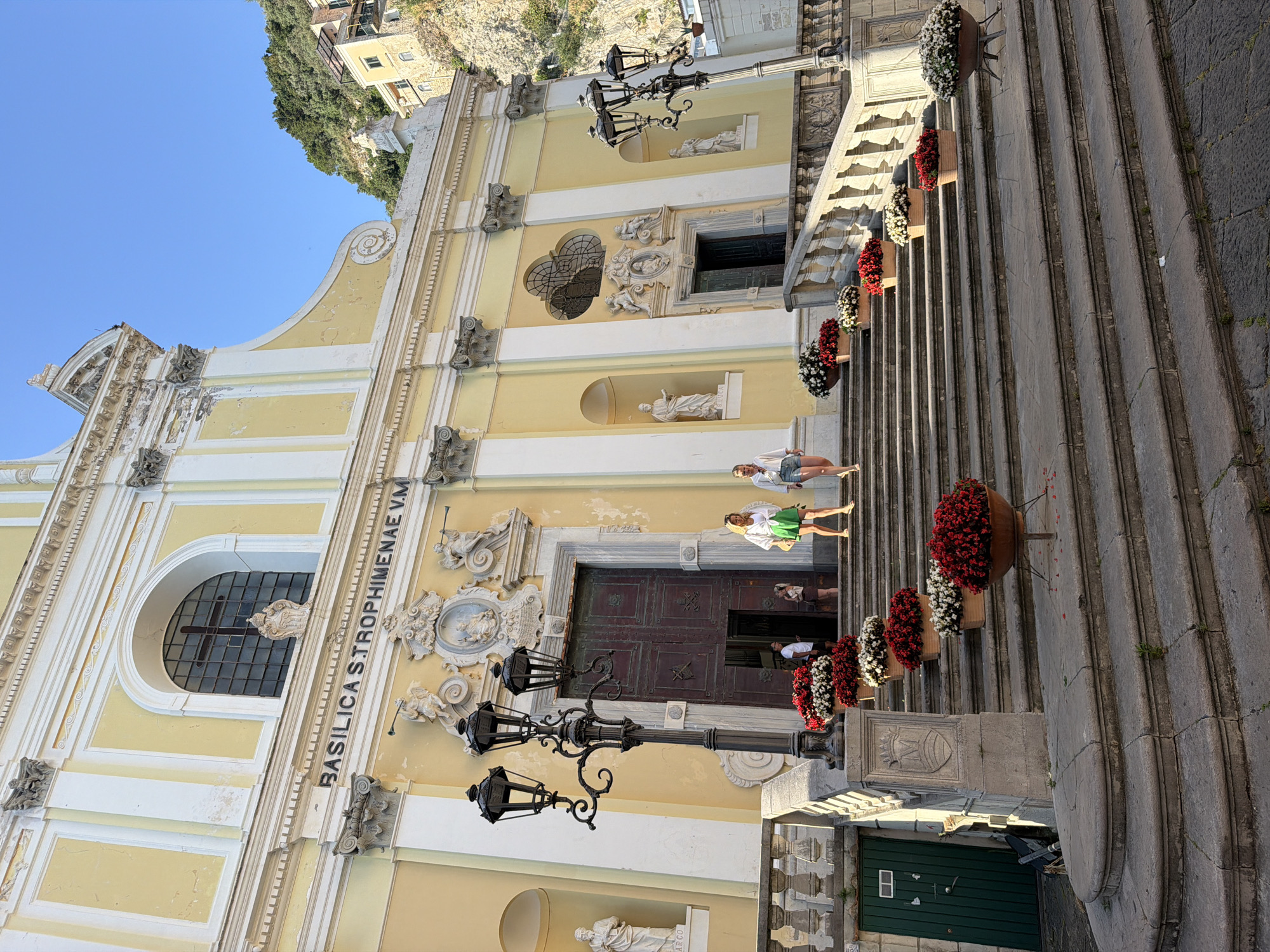 The image depicts the Basilica di Santa Trofimena, located in the city of Minori, Italy. The basilica has a yellow facade and white trim, with a large arched window and the inscription "BASILICA STROPHIMENAE V.M." on its left side. There are several statues in niches along the facade. A large set of stairs leads up to the main entrance, decorated with flower pots filled with red and white flowers. Two women are walking down the steps. A lamp post stands near the base of the stairs. The sky is blue.