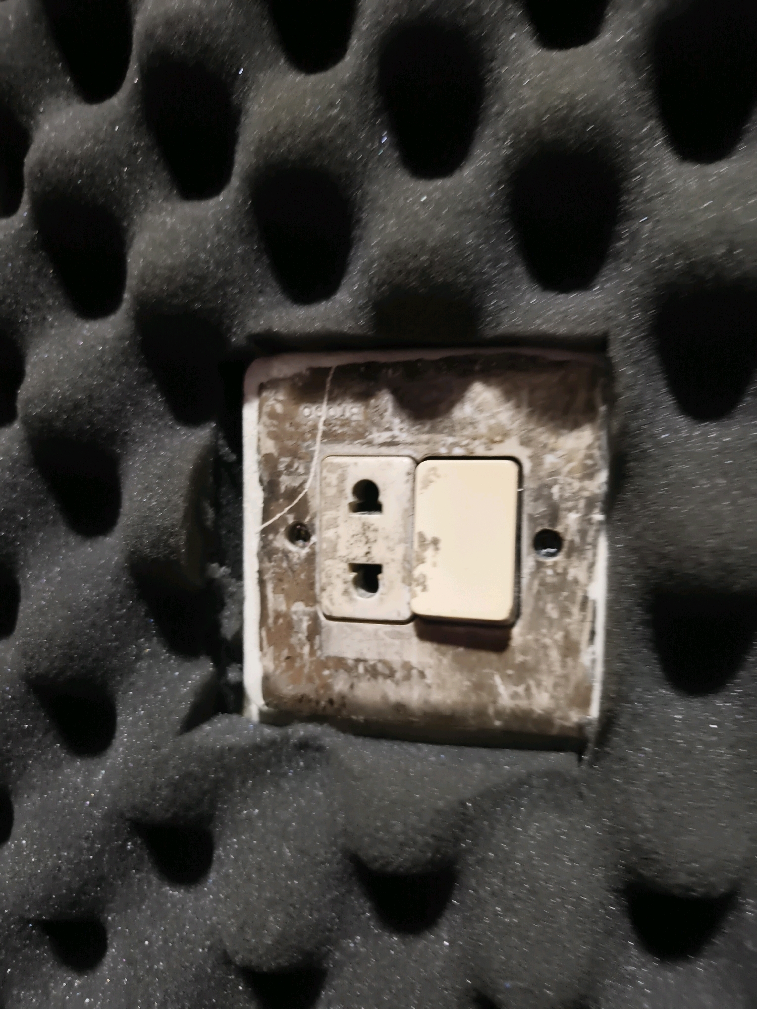An electrical outlet with a light switch is mounted on a textured black acoustic foam wall. The outlet and switch plate are discolored and appear aged, with some visible smudges and a thin white string draped across it. The surrounding acoustic foam has a dimpled pattern, typical of soundproofing material. The scene is likely an indoor environment. It is not possible to determine the specific location in Cilegon, Indonesia, or the broader context of the image from the visual information provided.