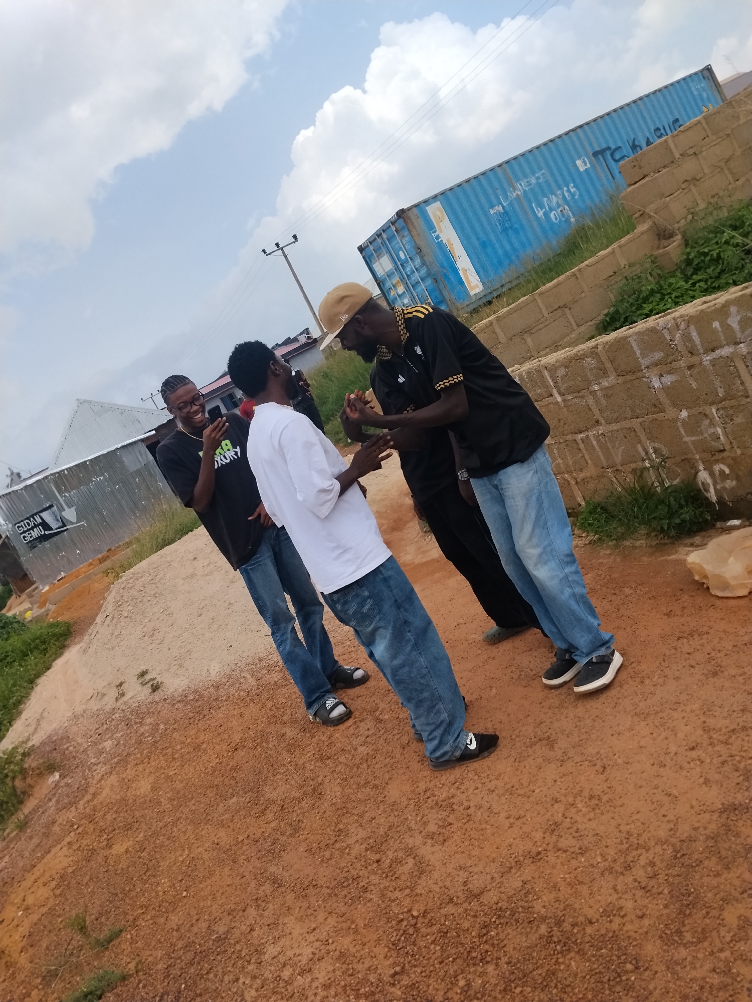 The image depicts a scene in the city of Durumi, Nigeria, featuring four individuals in an outdoor setting. Two men, one wearing a black t-shirt with glasses and the other in a white t-shirt, are engaged in conversation, with the man in the black t-shirt holding a phone. A third man, in a black shirt with yellow trim and a beige baseball cap, stands facing them, also interacting. A fourth person, partially obscured, is visible in the background. The environment is characterized by a dirt ground, a corrugated metal building with signage, a brick wall, and a large blue shipping container in the background. The sky above is partly cloudy. The overall impression is of a casual social gathering in an urban or semi-urban area.