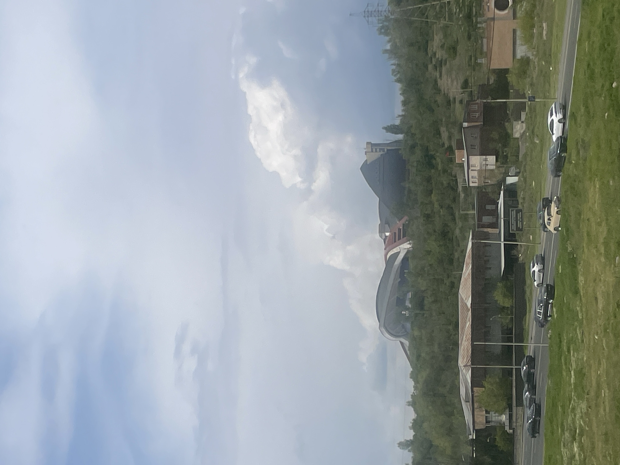 This image captures a daytime scene of an urban landscape in Yerevan, Armenia, likely from an elevated perspective looking down and across. The weather appears partly cloudy with large, soft white and grey clouds dominating the sky.
On the right side of the frame, a paved road stretches into the distance, with several vehicles visible. These include a white SUV, a dark sedan, a light-colored car (possibly beige or champagne), and a line of dark-colored cars further ahead, suggesting active traffic. The road is bordered by wide, lush green grassy areas.
To the left of the road, a mix of buildings and natural elements are present. A prominent feature is a large, modern building with a distinct curved roof and facade, displaying grey and reddish-brown sections. This structure is identifiable as the Karen Demirchyan Complex, a significant landmark in Yerevan. Behind and slightly to the right of this complex, another large, dark, blocky building stands, possibly an industrial or commercial structure. Further along the roadside, closer to the foreground, are several smaller, older buildings, one with a distinct rusty corrugated metal roof, nestled among dense green trees and vegetation. In the distant upper-left, power lines and transmission towers are visible against the sky.
The scene conveys a sense of a typical day in a city, with regular traffic flow and a blend of modern and older architectural styles amidst a verdant landscape. No discernible text is visible on any signs, vehicles, or buildings.