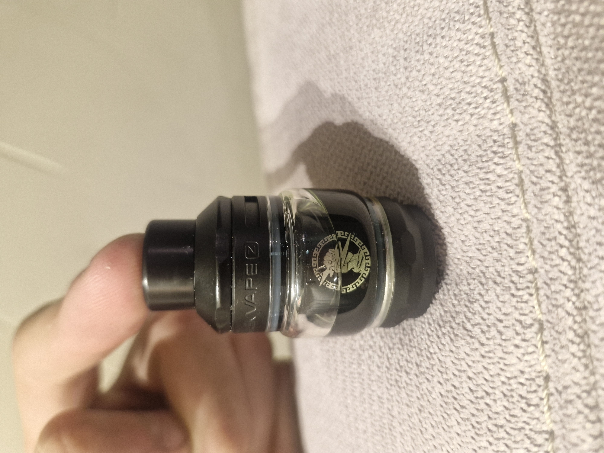 The image shows a black and clear vape tank, possibly a Zeus brand, held by a human finger. The tank is resting on a textured, light-colored surface. The environment appears to be indoors with artificial lighting. There is no information to suggest a specific location in Sofia, Bulgaria.