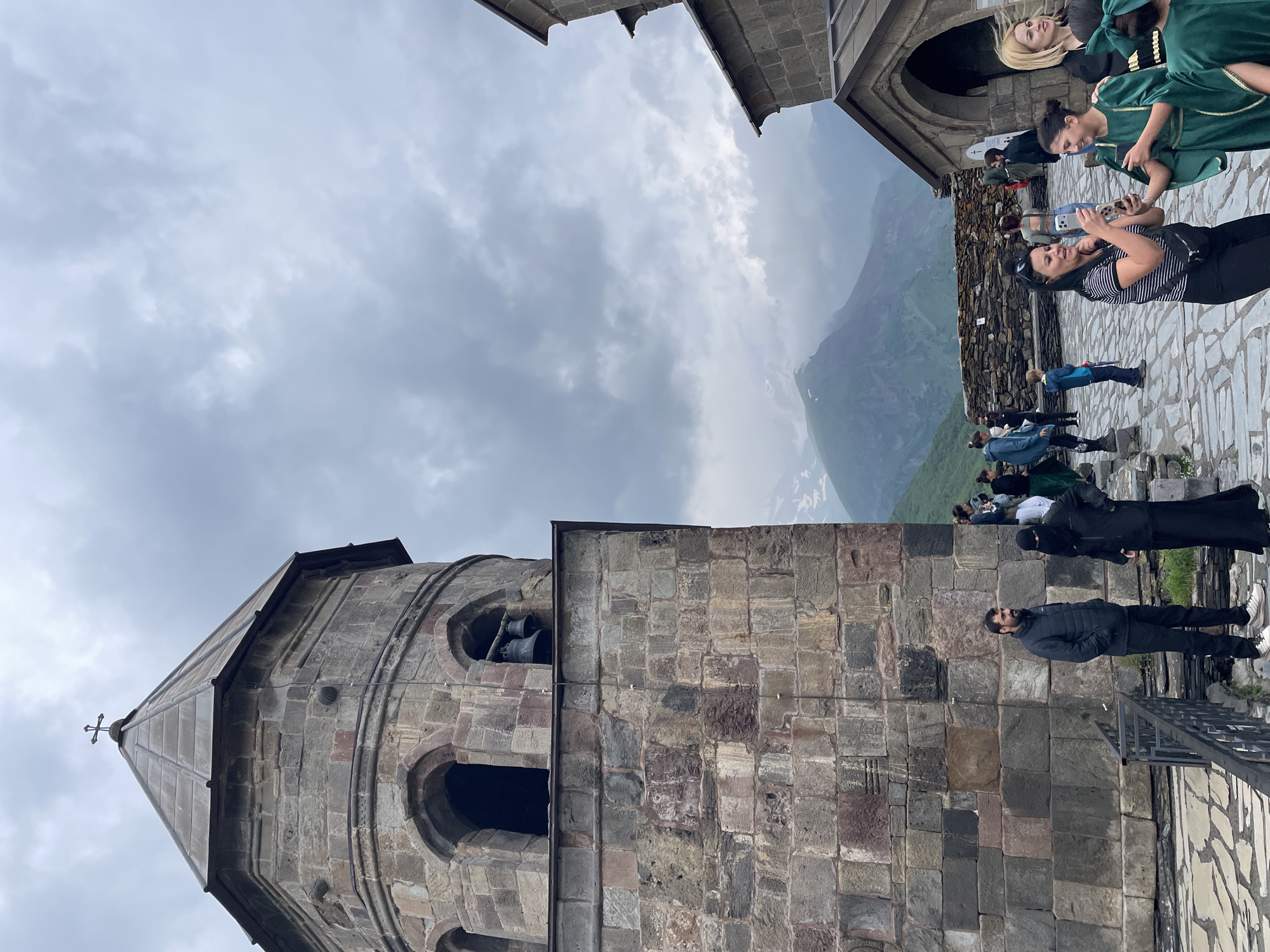 In Stepantsminda, Georgia, Gergeti Trinity Church stands prominently on a rocky elevation. The stone structure features a tall, octagonal bell tower with arched openings and two bells hanging within. The church itself is constructed from rough-hewn stones, with a sloped, metal-covered roof. A cross adorns the apex of the bell tower.
In the foreground and midground, a group of tourists are gathered on a paved stone courtyard. Several individuals are dressed in traditional green garments, including a young girl holding a smartphone and smiling. Another woman in a striped shirt is also taking a photo with her phone. Two people are standing near a black metal railing, one a man in a dark jacket and the other a woman in black attire, including a niqab. Other tourists are visible in the background, including a person in a blue jacket, a child in a blue sweatshirt, and others observing the scenery.
Behind the church, a dramatic mountainous landscape unfolds under a cloudy sky. The mountains are covered in green vegetation and snow-capped peaks are visible in the distance. A dry stone wall runs horizontally across the midground, separating the church area from the mountain slopes. The overall scene depicts a blend of historical architecture, cultural attire, and the breathtaking natural beauty of the Georgian Caucasus region.