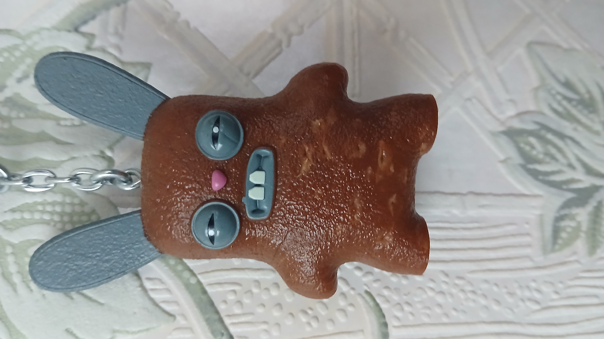 The image depicts a brown, toy-like figure of a rabbit on a light-colored surface. The rabbit has grey ears, eyes, and what appears to be a stylized mouth with two white teeth. A silver chain is attached to the back of the rabbit's head. The object is situated against a patterned background.