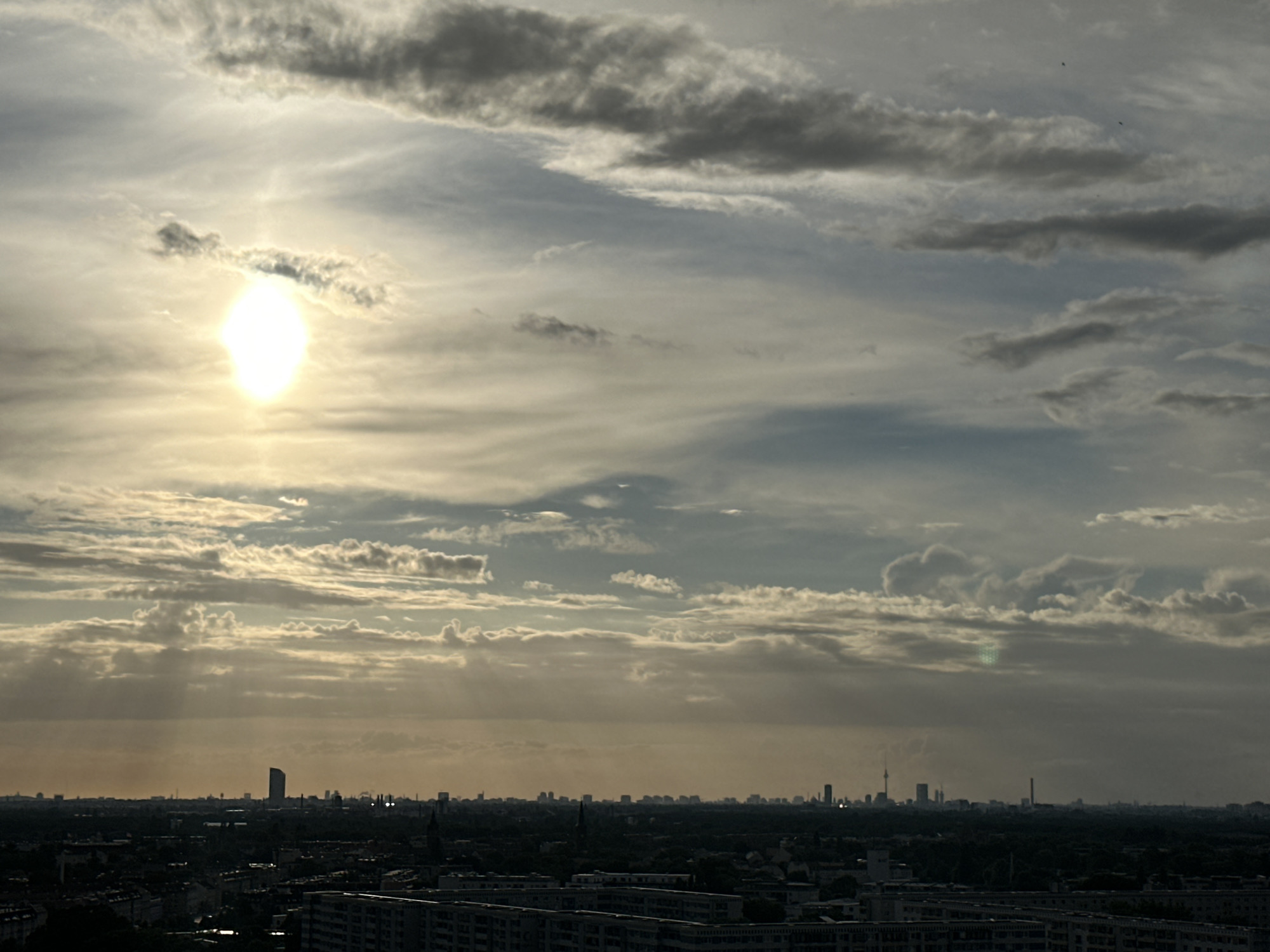 This is a high-angle view of the Berlin skyline at sunset, with the sun prominently positioned in the upper left quadrant, partially obscured by clouds. The sky is filled with a mix of dark and light grey clouds, with rays of sunlight piercing through in some areas. The city itself is silhouetted against the hazy horizon, with various buildings, including the distinctive TV Tower (Fernsehturm) visible in the distance. The foreground shows a densely populated residential area with apartment buildings. The overall impression is of a dramatic and somewhat moody urban landscape at dusk.