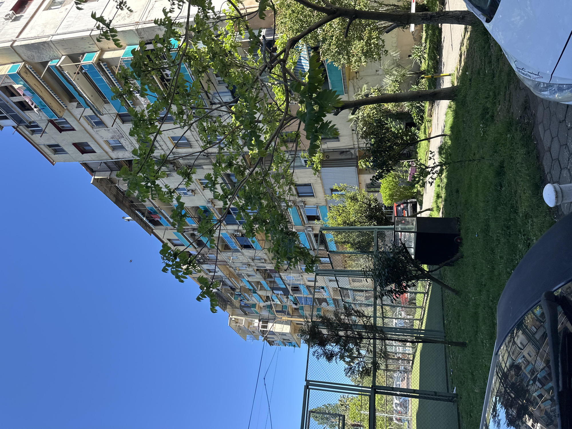 The image depicts a multi-story building, typical of Soviet-era architecture, in Batumi, Georgia. The building facade is off-white with rows of windows and protruding balconies, many of which are shaded with blue awnings. The balconies appear weathered and some have items hanging from them.
In the foreground, a metal fence runs along the edge of a grassy area. A few trees are visible, their branches partially obscuring the building. Adjacent to the grass is a paved walkway. Parts of vehicles, likely parked cars, are visible on both sides of the image. The sky is clear blue, suggesting a sunny day.