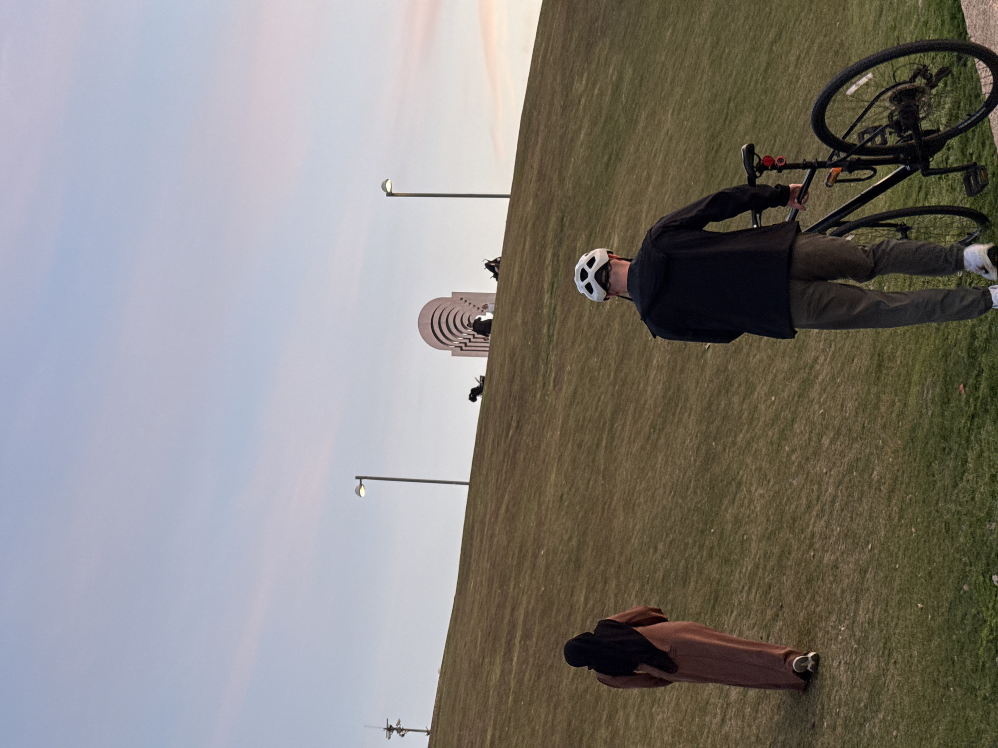 A man stands beside a bicycle atop a grassy hill in Tel Aviv, Israel. The location is likely Park Haherez. In the background is the Rabin Memorial, which is a series of arches. A woman is bent over a short distance away. Streetlights are visible in the background. The sky is pale with faint wisps of pink.