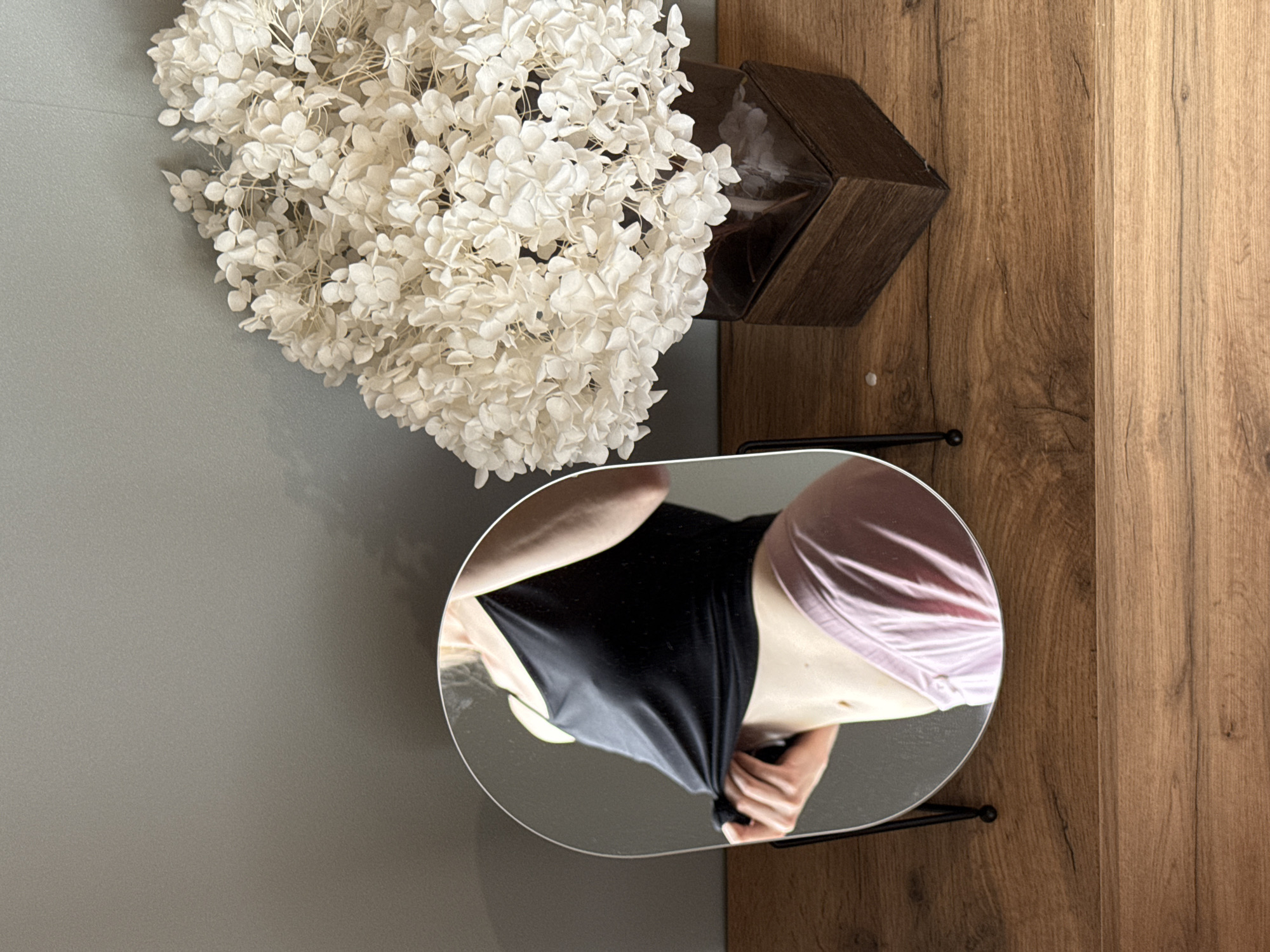 The image shows a scene inside an indoor space, possibly in Chaikovsky, Russia, given the prompt's stipulation. The view is from an elevated angle, showcasing a portion of a room with three main elements: a floral arrangement, a wooden container, and a mirror reflecting a person.
The floral arrangement, consisting of white flowers (likely hydrangeas), is located in the upper left quadrant of the image. To its right, positioned on a wooden surface, is a dark-colored, geometric wooden container.
Below these elements, a circular or oval mirror is present, reflecting a person wearing a black top and pink shorts or undergarments. The person's hand is visible, appearing to adjust the black top. The mirror is situated near the edge where the gray wall meets the wooden surface.
