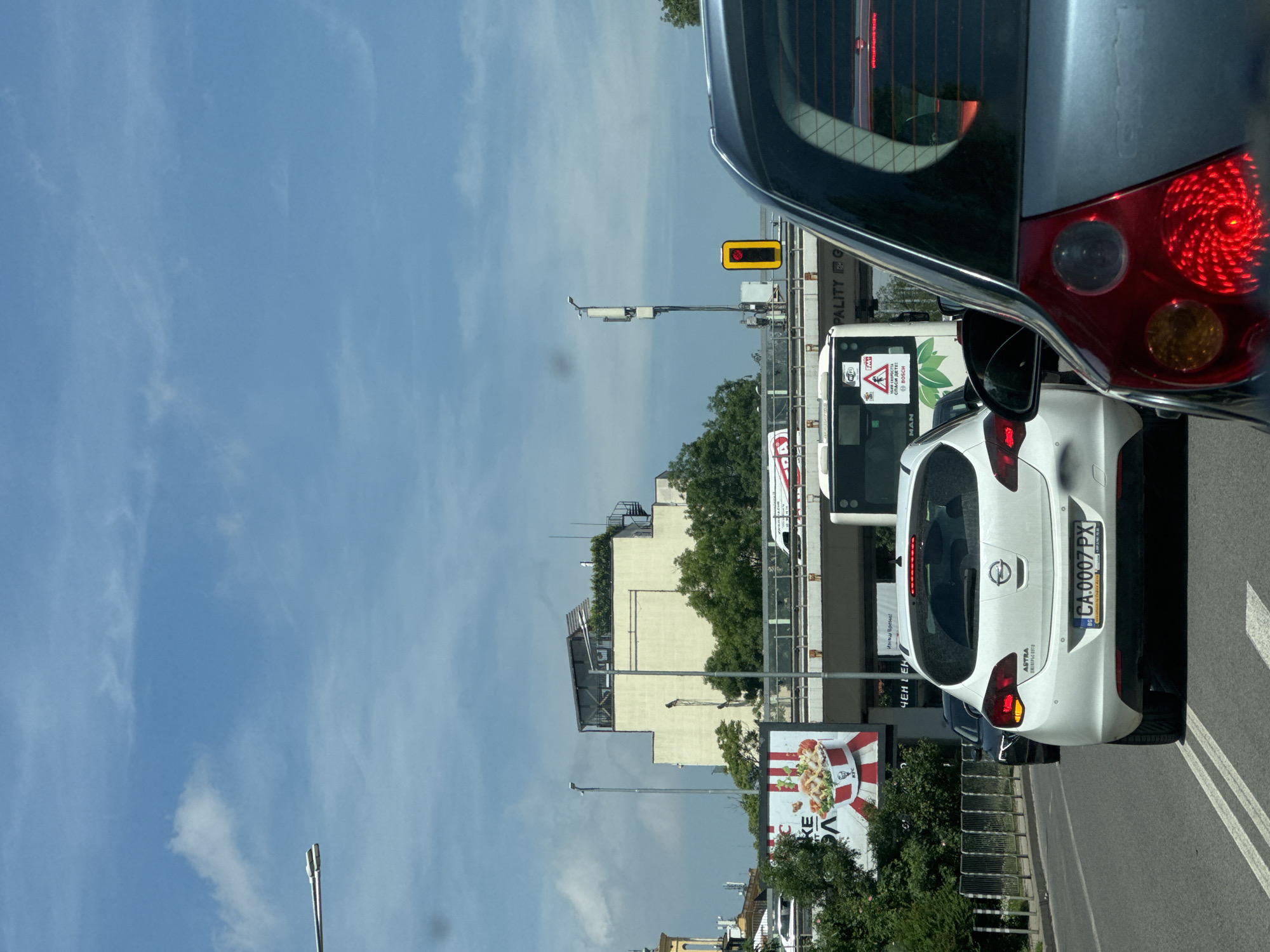 This image captures an urban traffic scene from the perspective of a vehicle, looking forward and slightly to the right. The top-right portion of the frame shows the rear of the car from which the photo is taken, including a red tail light and dark grey bodywork.
Ahead, a white Opel Astra hatchback is prominently featured, displaying its Bulgarian license plate `CA.0007 PX`. The text "ASTRA" and "ЕЛЕЛЕРЬС АУТО" are visible on its rear. Behind the Opel and further to the left, a white bus is seen, bearing several signs including "Wi-Fi," a green leaf symbol, "СПАСИ ДЕТЕ!" (Save the Child!), "BOSCH," and "MAN." The vehicles appear to be on a multi-lane road, indicated by white markings on the asphalt.
In the background, a light-colored building with some trees on its roof is visible. A large billboard with "KE" and part of a KFC bucket meal is partially obscured by trees in the lower left. Above the bus, a traffic light with a red illuminated signal in a yellow casing is mounted on a pole. Further in the background, a structural element, possibly an overpass or elevated platform, displays partially visible text "PALITY G" (likely part of "Municipality"). The sky is bright blue with scattered thin white clouds, suggesting clear daytime weather. The scene depicts typical city traffic in Sofia, Bulgaria.