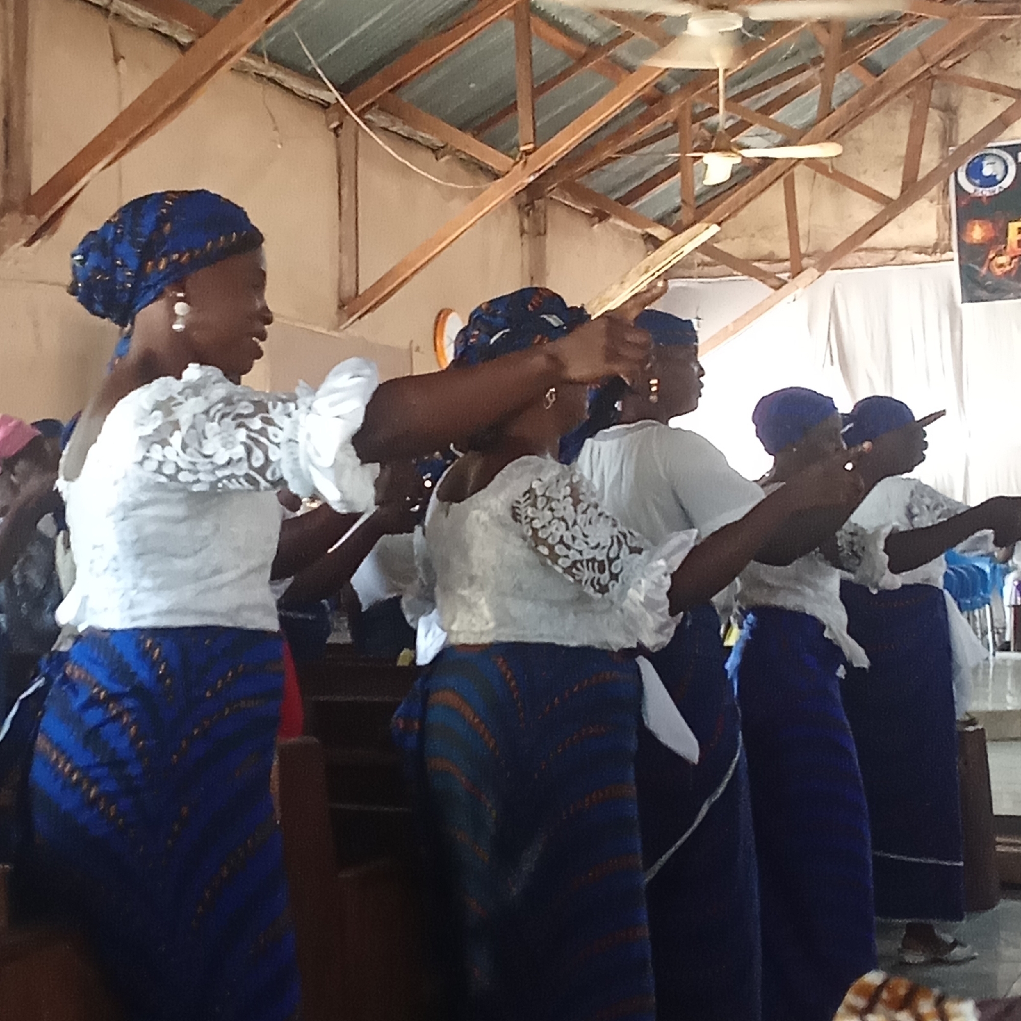 A group of women are performing a synchronized dance in what appears to be a church setting in Unknown, Unknown. They are dressed in matching outfits consisting of a white top with lace details, a blue patterned skirt, and a blue headwrap. The women are standing in a row, extending their arms forward in unison. The location features a simple interior with wooden beams supporting the ceiling and a ceiling fan visible. There is a poster on a wall, and the presence of pews suggests a religious context.