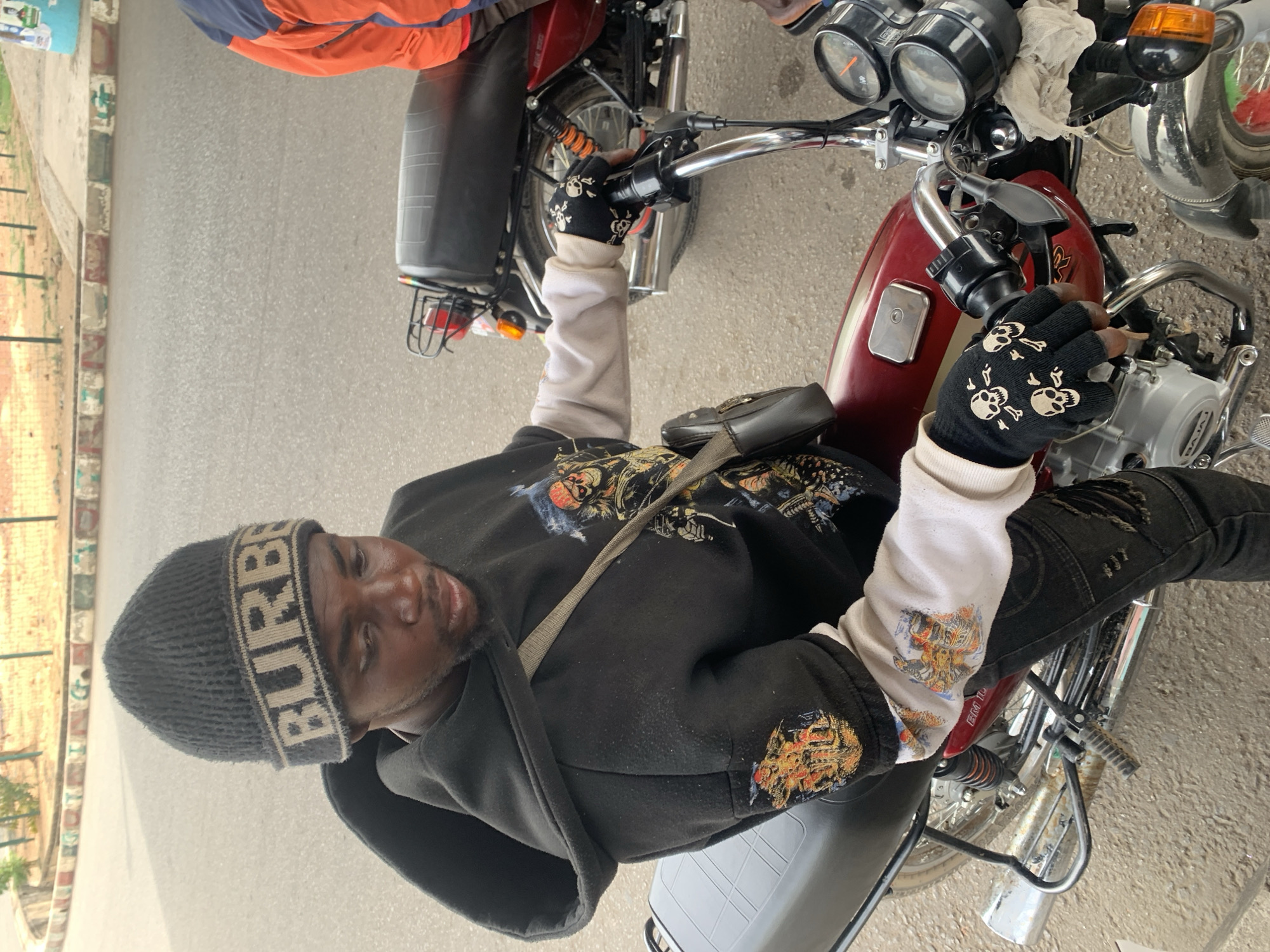 A man sits on a parked motorcycle in Jos, Nigeria. He wears a black beanie with "BURBERRY" on it, a black hooded jacket over a long-sleeved white shirt with patches, black distressed jeans, and black fingerless gloves with skull designs. He has a small black bag slung across his chest. The motorcycle is maroon and silver. The road and curb are visible in the background.