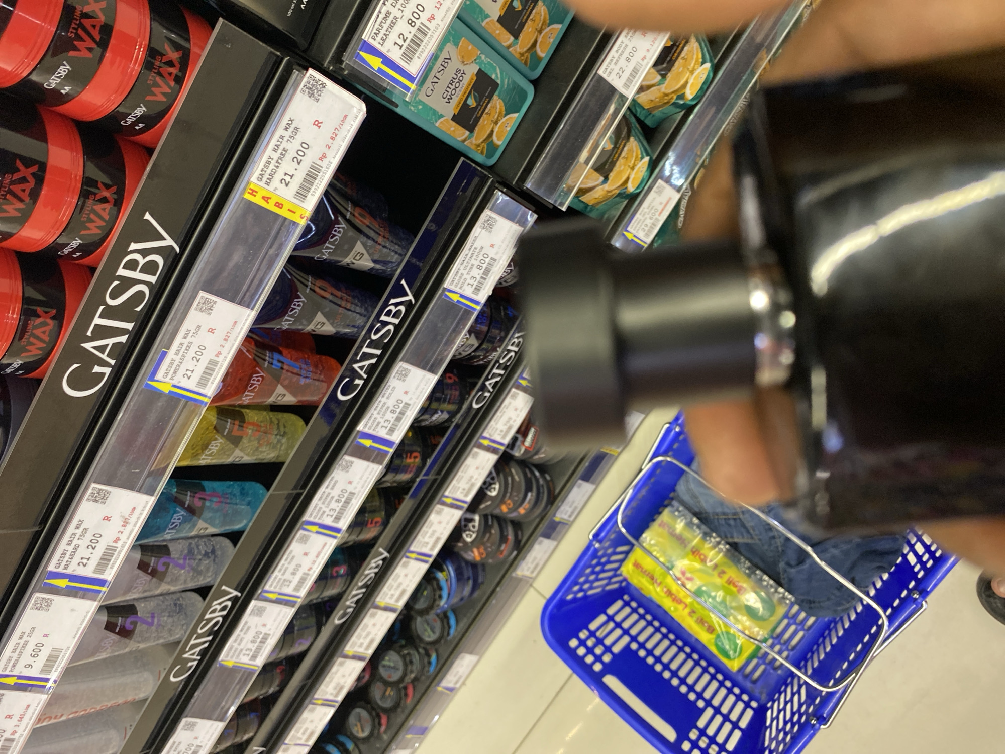 The image shows a retail store aisle with shelves stocked with Gatsby hair styling products. The products include hair wax in various types like "Power & Spikes," "Hard & Free," and "Glossy Ultimate." There are also hair gels and "Citrus Woody" hair care products visible. The pricing tags indicate prices in Indonesian Rupiah. In the foreground, a blue shopping basket contains some items, including what appears to be jeans and a yellow package. A person's hand, holding a black bottle, is partially visible, obscuring some of the products. The overall environment suggests a well-lit supermarket or convenience store. The presence of Indonesian Rupiah and product names commonly found in the Indonesian market suggests the location is in Indonesia, and given the context of previous interactions, the city is likely Banda Aceh.