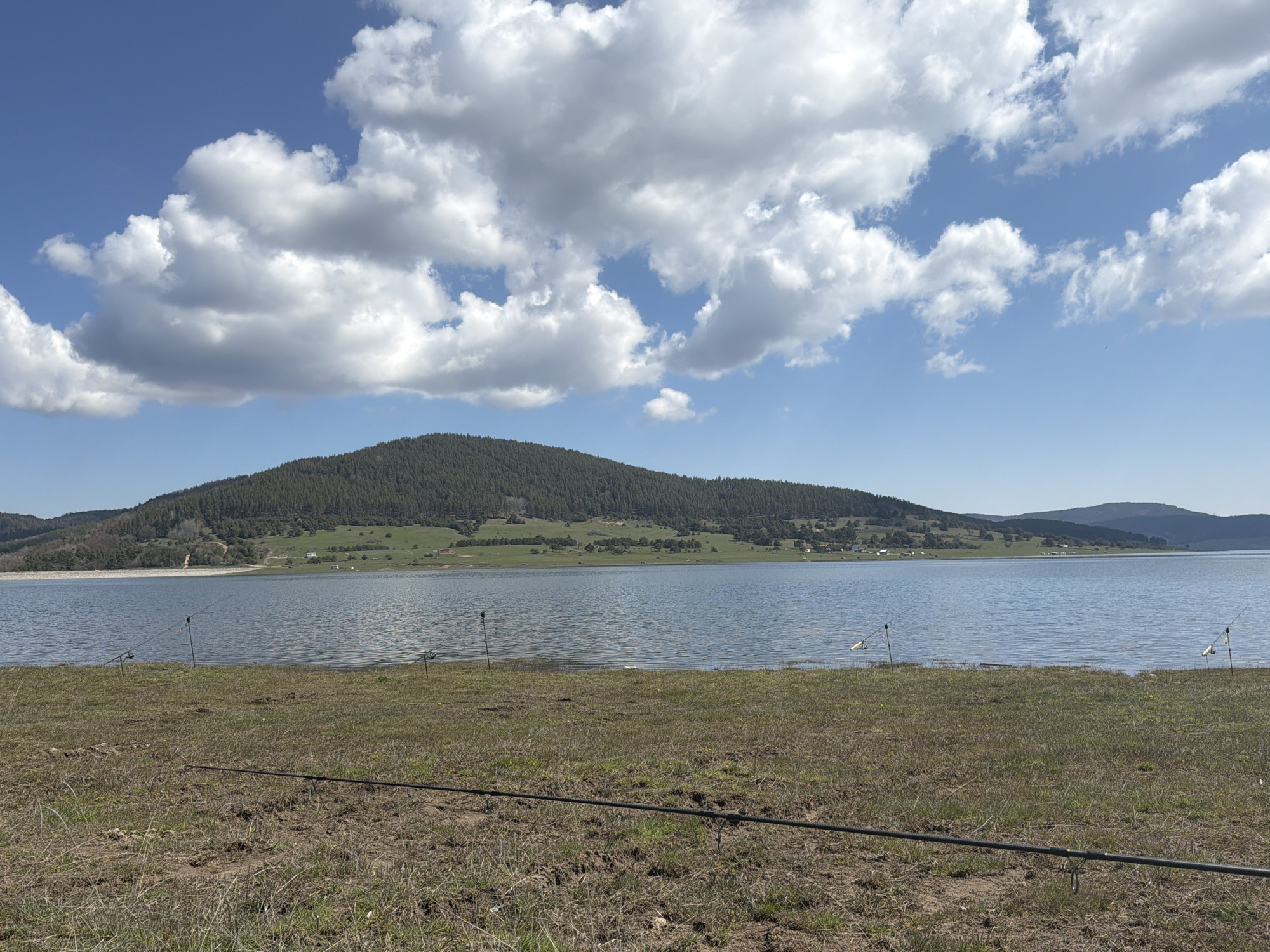 The image shows a scenic view of Batak Reservoir near Rakitovo, Bulgaria. The foreground features a grassy bank with fishing rods laid out. The water of the reservoir extends to the middle ground, with a forested hill in the background. The sky is blue with fluffy white clouds.