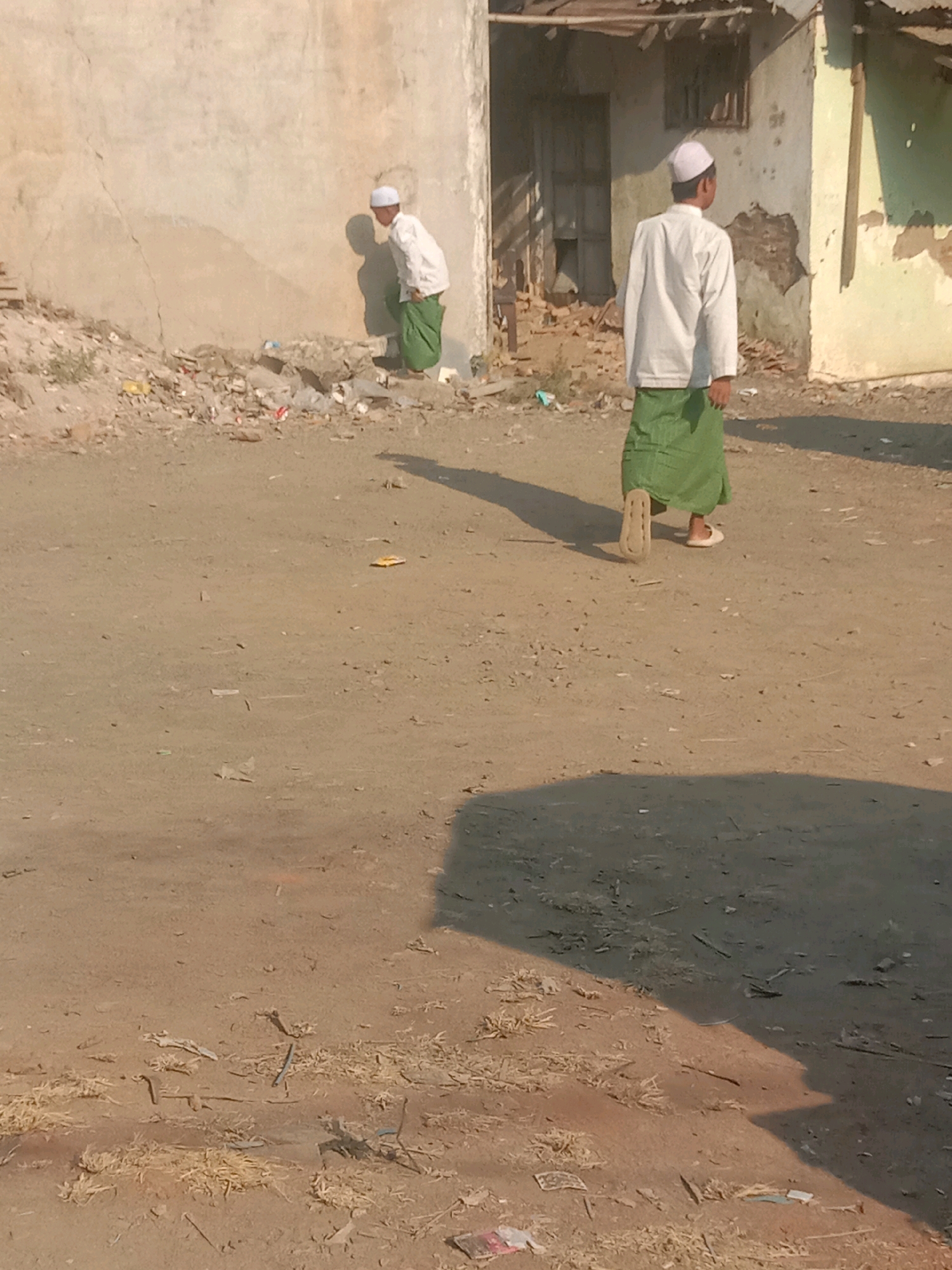 The image depicts an outdoor scene in Sidogiri, Indonesia, on a dirt ground with scattered debris. In the foreground and middle ground, two individuals, likely male, are present. The person on the left, appearing to be a child, is wearing a white long-sleeved shirt and a green sarong, with a white cap. This individual is bending over, facing a rough, textured wall. Behind this person, there is a shadow cast on the wall and ground.
Further into the middle ground, another person is seen from the back, also wearing a white long-sleeved shirt, a green sarong, and a white cap. This individual is walking away from the viewer towards a building. The building in the background appears weathered, with damaged walls and a corrugated metal roof. The ground is uneven and sandy, with visible bits of straw and other discarded materials. Shadows are prominent, indicating bright sunlight. The overall environment suggests a rural or semi-urban setting with visible signs of disrepair and neglect.