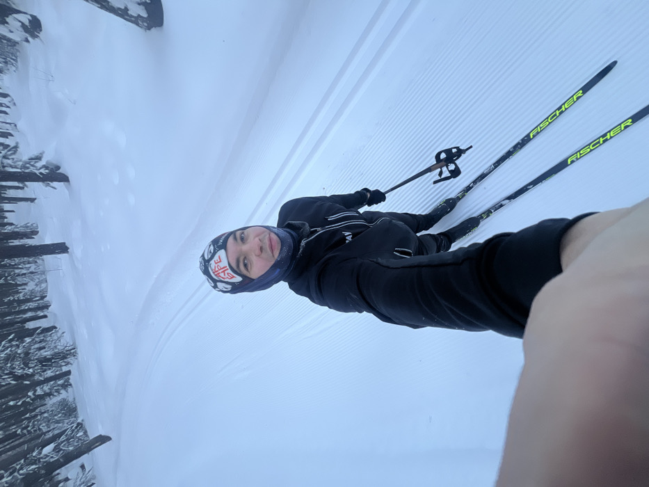 An individual is depicted cross-country skiing on a groomed trail within a snow-covered forest in Kazan, Russia. The male subject, positioned centrally and looking towards the camera, appears to be taking a selfie with his right arm extended. He wears a multi-colored beanie with visible "АК БАРС" text, a dark balaclava/neck gaiter with frost around the mouth, and a black athletic jacket with a partially visible "SAV" logo. His left hand grips a ski pole. He stands on two Fischer-brand cross-country skis, which are black with green/yellow highlights, aligned within machine-groomed classic ski tracks. The surrounding environment is a dense coniferous forest, with trees heavily laden with white snow. The ground is entirely snow-covered, displaying both classic ski tracks and a wider, ribbed skate skiing surface. The lighting is diffuse, indicative of an overcast winter day.