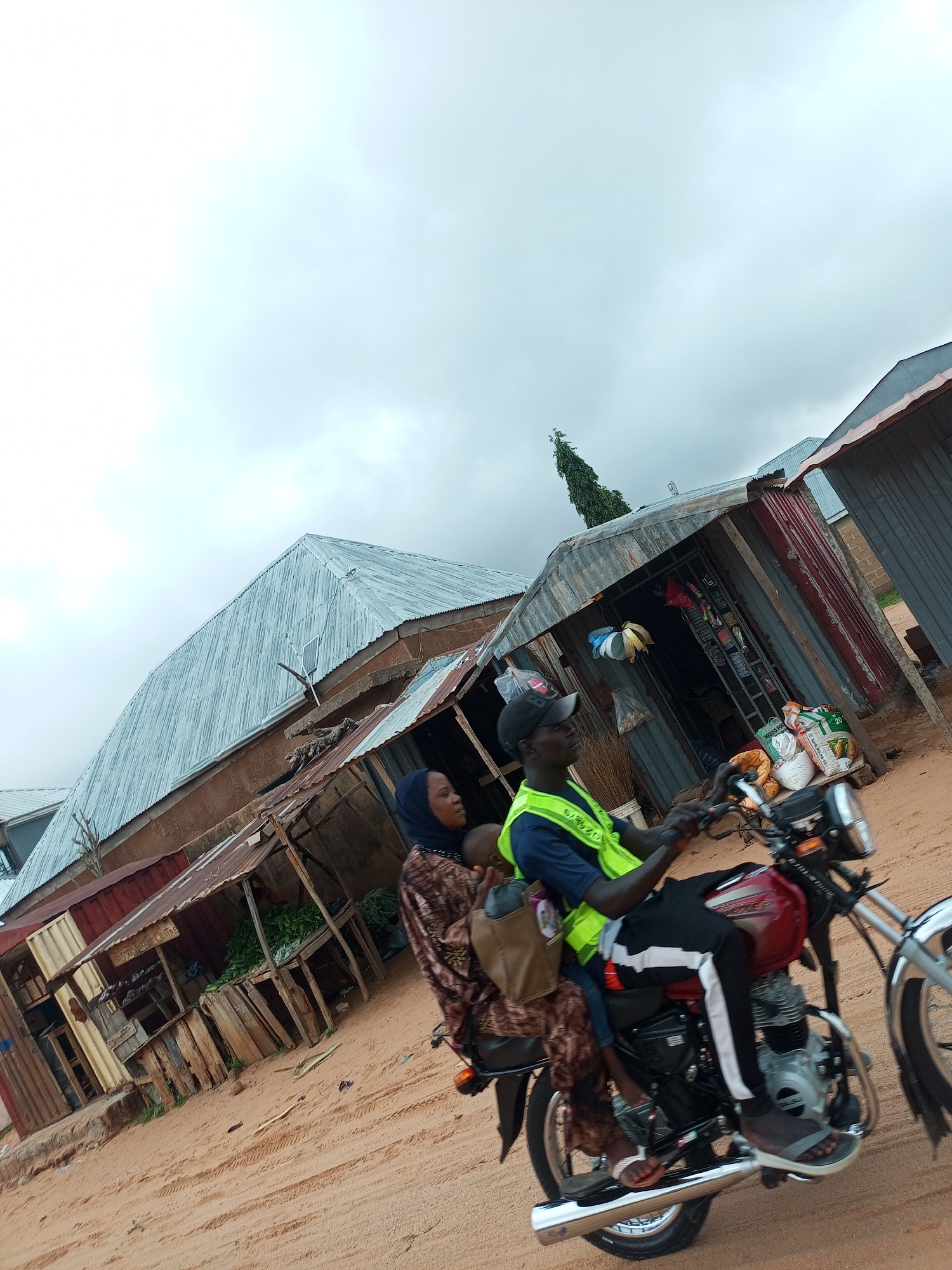 The image depicts a street scene in Tempure, Nigeria, on an overcast day. A man wearing a neon yellow vest and a black cap is riding a red motorcycle. He is carrying a woman behind him, who is wearing a patterned brown and white dress and a blue headscarf. The woman is holding a baby. The motorcycle is in motion, suggesting they are traveling.
In the background, there are several market stalls with corrugated metal roofs. Some stalls appear to be selling goods, with items like bags of fertilizer and produce visible. The structures are weathered and made of various materials, including corrugated metal and wood. The ground is sandy, with tire tracks visible, indicating a dirt road or unpaved area. A tall, thin tree is visible in the distance, with a slightly conical shape. The overall environment suggests a rural or semi-urban setting within Tempure.