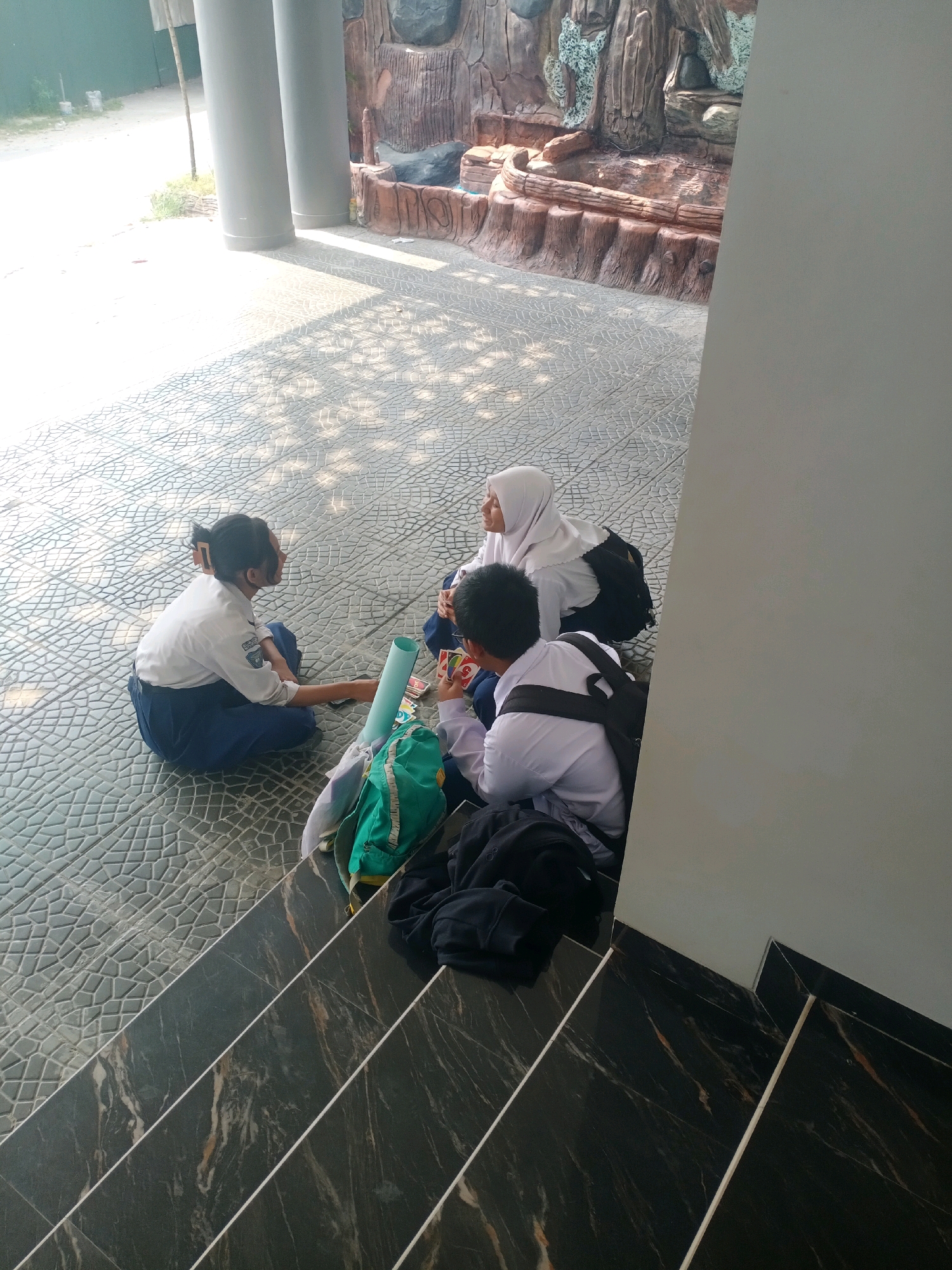 Three students in school uniforms are sitting on a patterned tiled ground near a decorative rock wall and a building wall. The student on the left is wearing a white shirt and a dark blue skirt, with their hair tied back. They are holding a cell phone and a light blue tube. The student in the middle is wearing a white hijab and a white shirt, with a backpack. The student on the right is wearing a white shirt and dark pants, also with a backpack, and appears to be holding playing cards. Two backpacks, one green and one black, are on the ground near the students. The scene is outdoors, with sunlight casting shadows on the ground. The location is not specified within the image itself, but given the context of Samarinda, Indonesia, it suggests an outdoor school area or public space within the city.