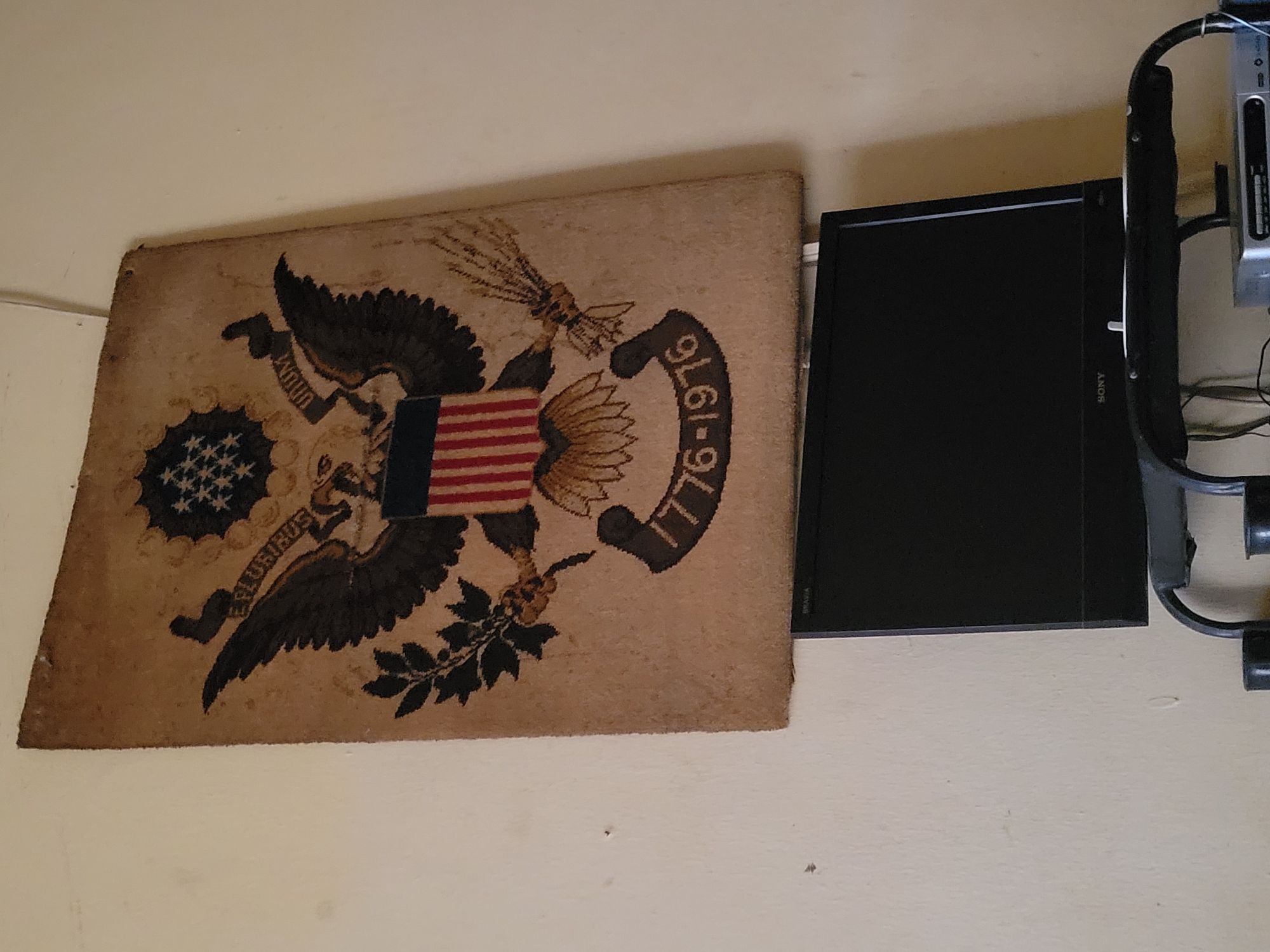 A tapestry featuring the Great Seal of the United States hangs on a wall next to a Sony Bravia flat-screen television. The tapestry is rectangular and shows a stylized eagle, stars, and stripes, along with the dates "1776-1976". The wall is light beige in color. Some cables and mounting hardware are visible behind the television. The city and location are unknown.