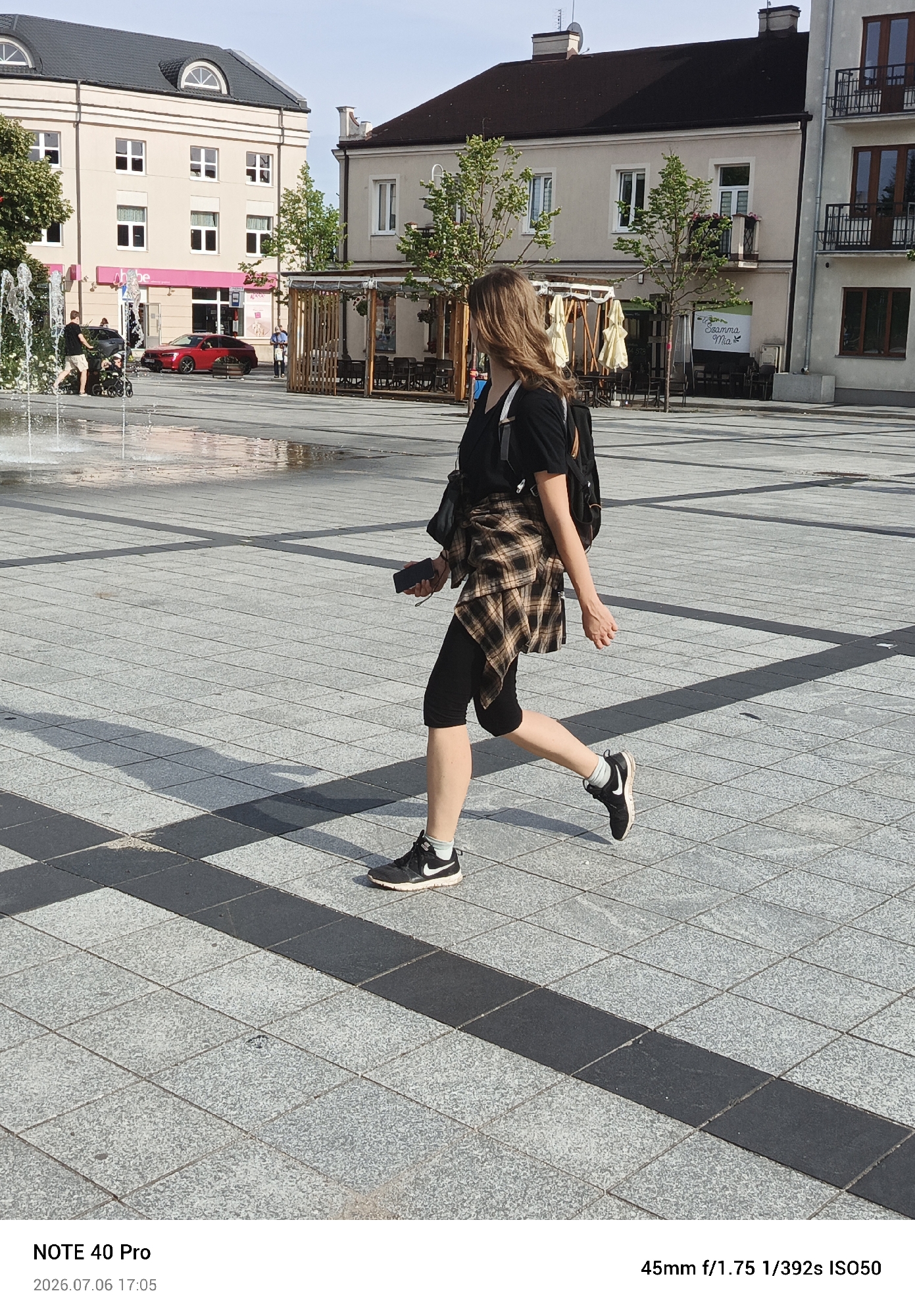 A woman is walking across a paved square in Piaseczno, Poland, on July 6, 2026, at 5:05 PM. She is wearing a black t-shirt, black leggings, black sneakers with white socks, and a plaid shirt tied around her waist. She has a black backpack on and is holding a black smartphone in her right hand. She is walking from left to right in the frame, with her left leg extended forward. The square is paved with large square tiles and has dark striped borders. In the background, there are buildings with windows and balconies, trees, an outdoor seating area with tables and chairs, and a fountain. A red car is parked to the left of the scene. The sky is blue with some clouds. The image was taken with a camera at a focal length of 45mm, aperture of f/1.75, shutter speed of 1/392s, and ISO of 50.