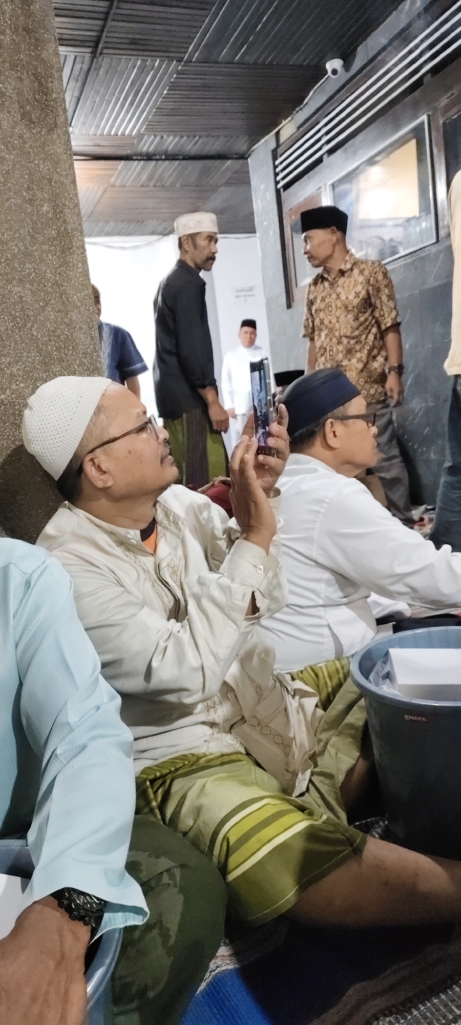 The image captures a gathering of men, likely in a mosque or community hall in Malang, Indonesia. In the foreground, a man wearing a white skullcap and glasses is holding up a smartphone, seemingly taking a photo or video. He is dressed in a cream-colored shirt with intricate embroidery and a green and white striped sarong. To his right, another man in a black skullcap and a patterned shirt is visible. In the background, several other men are present, some in traditional attire. The environment appears to be indoors, with a corrugated metal ceiling and textured walls. A security camera is mounted on the ceiling. The overall scene suggests a social or religious event.