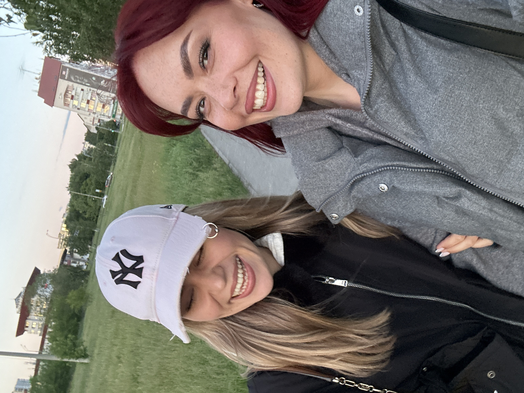 Two young women pose for a selfie outdoors. One woman has red hair and wears a gray jacket. The other woman wears a black jacket and a white baseball cap with the "NY" logo in black. Both are smiling broadly. The background includes a grassy area and buildings, consistent with an urban park setting. The location is Tolyatti, Russia, based on building design.