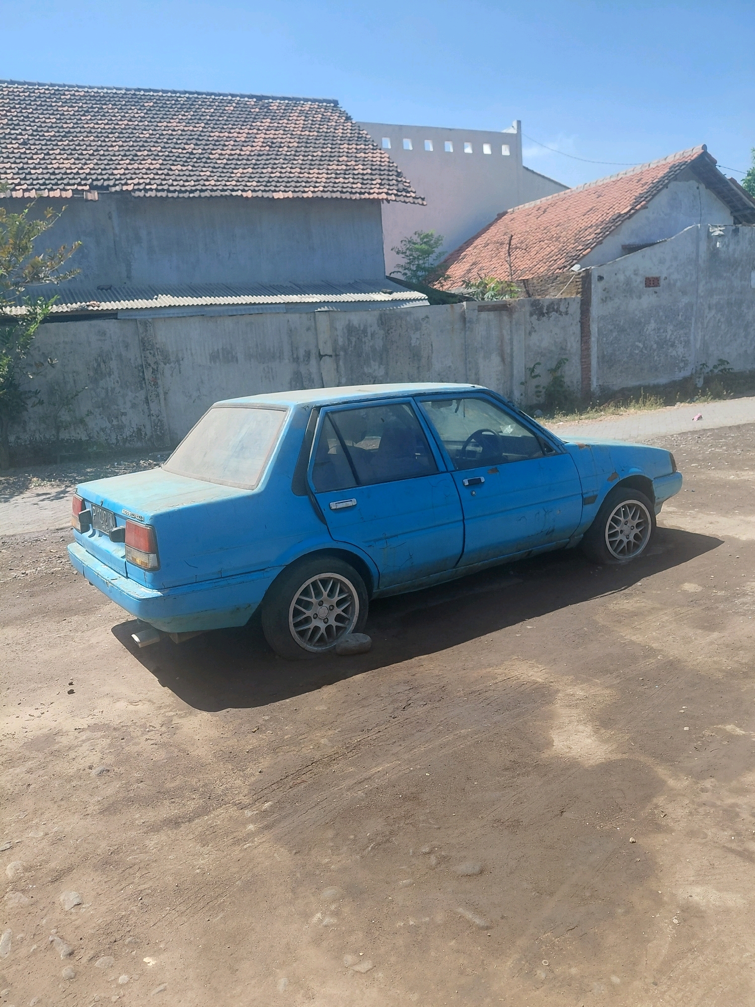 A dusty, bright blue sedan, possibly a Toyota Corolla, is parked on a dirt road in Paiton, Indonesia. The car appears to be in disrepair, with visible rust and chipped paint. The rear left tire is flat, and a rock is placed beneath the car to support it. The background features two houses with red tile roofs and a gray concrete wall. The sky is clear and blue, indicating a sunny day. The overall impression is one of abandonment or neglect in a rural or semi-rural setting.