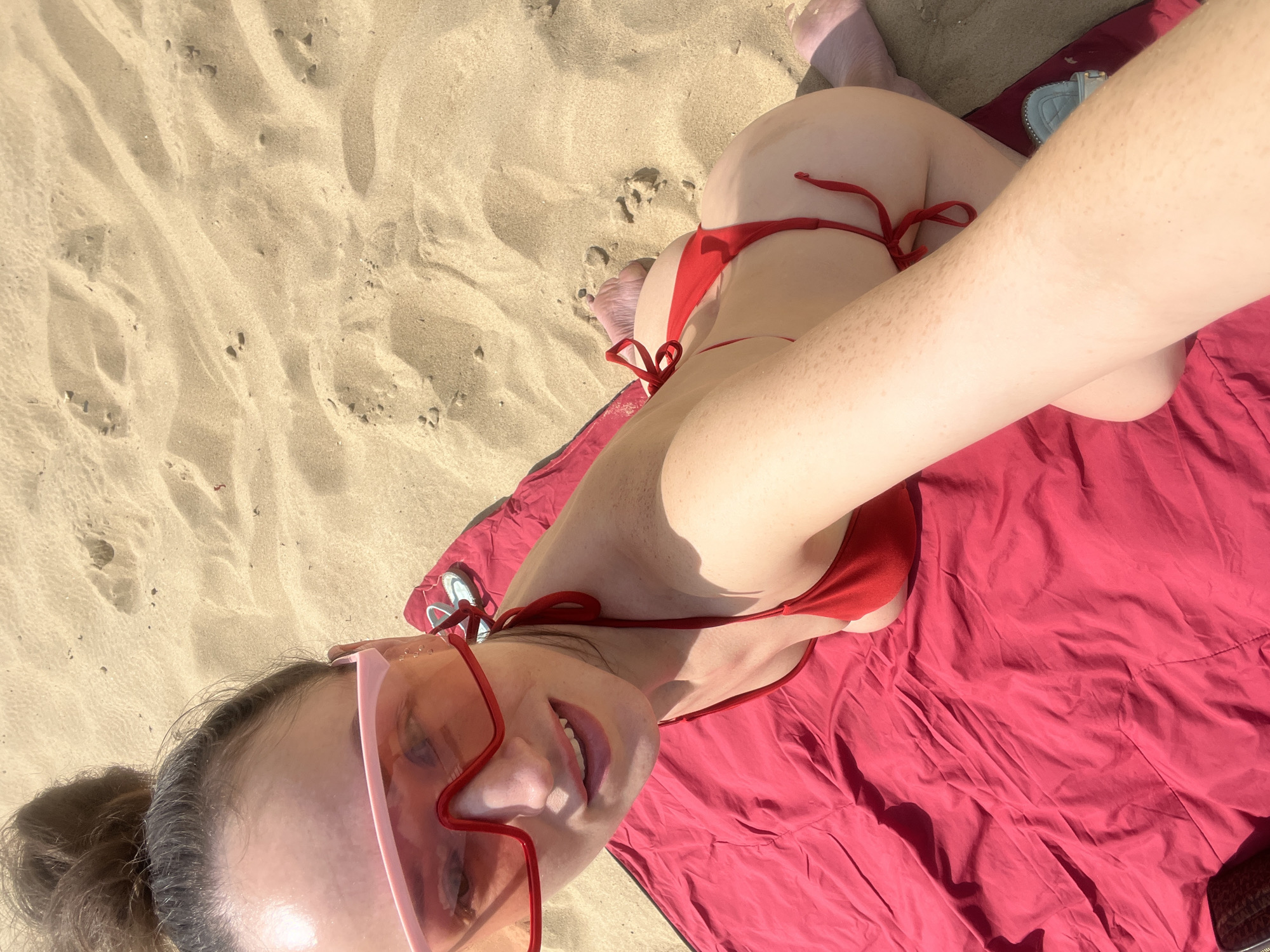 A person is seen from a high angle, lying on a red blanket on a sandy beach. The person is wearing a red bikini and large, pink, star-shaped sunglasses. The person is smiling and looking at the camera, holding it to take a selfie. The background consists of sand and a hint of water. The location is not specified as Kazan, Russia.