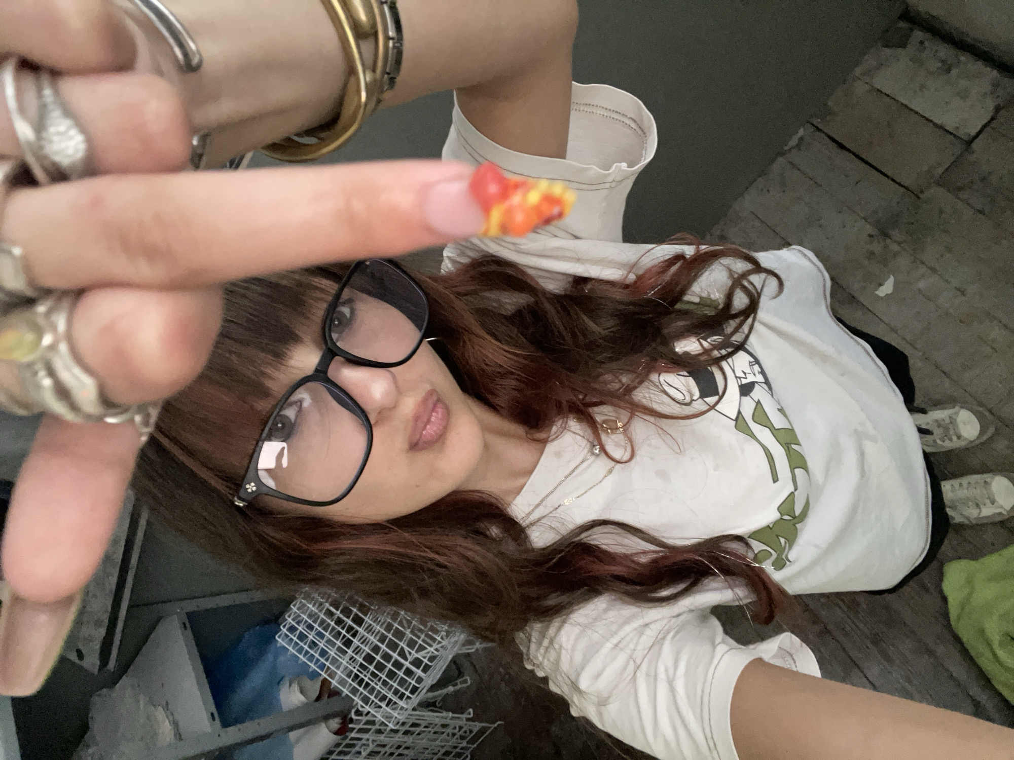 A young woman with long, dark, wavy hair, bangs, and glasses is shown from the chest up, taking a selfie. She is wearing a white t-shirt with a cartoon graphic and several necklaces. She is wearing rings on her fingers, and her right index finger is extended towards the camera, showing a brightly colored, patterned fingernail. She is wearing large, black-rimmed glasses. Her lips are slightly pursed. The background appears to be an indoor area with shelving units and some metal cages. The floor is made of wooden planks. The lighting is dim. The location is identified as Raanana, Israel.