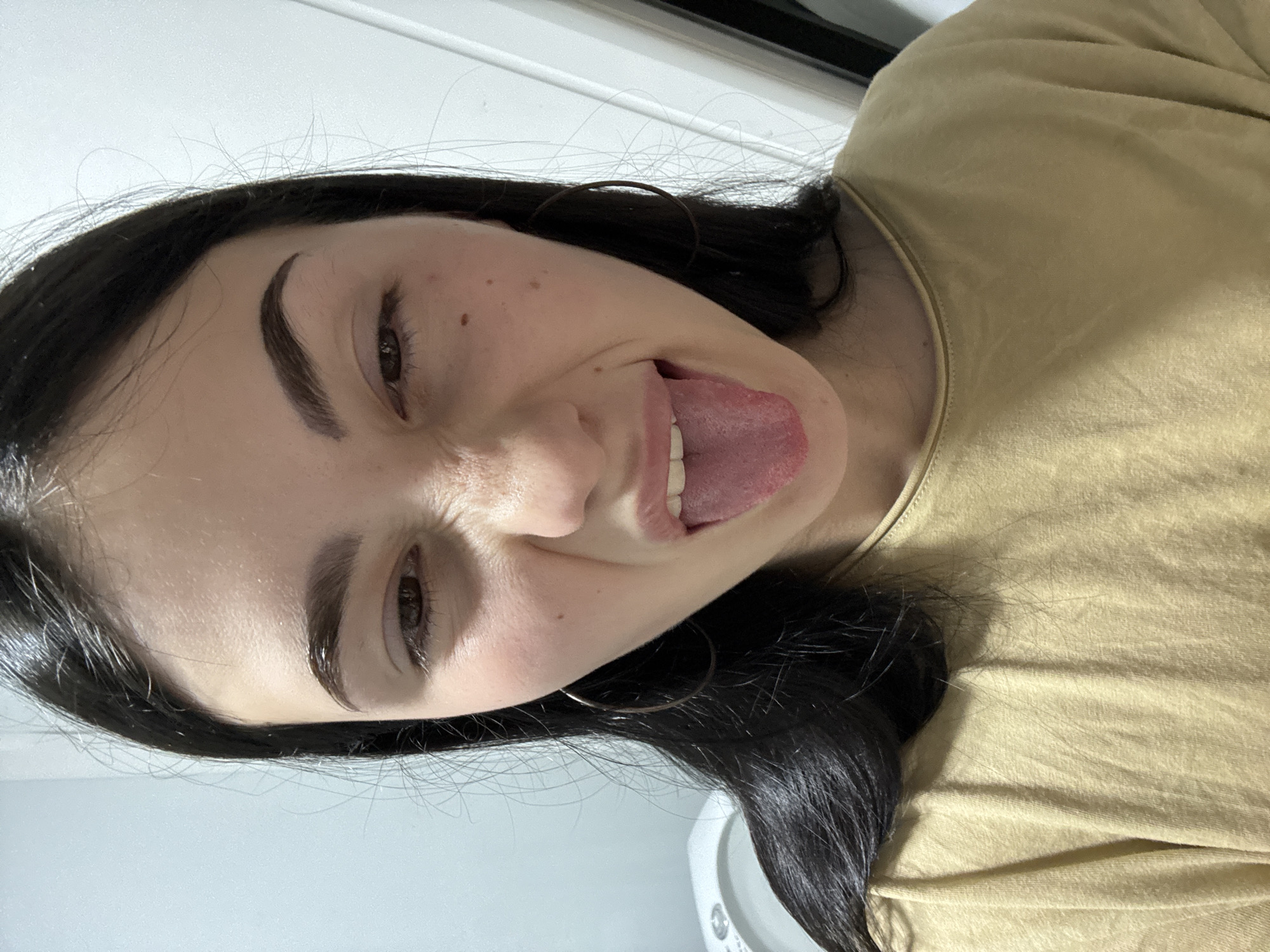 A young woman with dark hair and hoop earrings makes a face at the camera, sticking out her tongue. She is wearing a mustard-colored t-shirt. The background is a plain wall and what appears to be the top of a toilet tank, suggesting the photo was taken in a bathroom. The location is the city of Фоки, Russia.