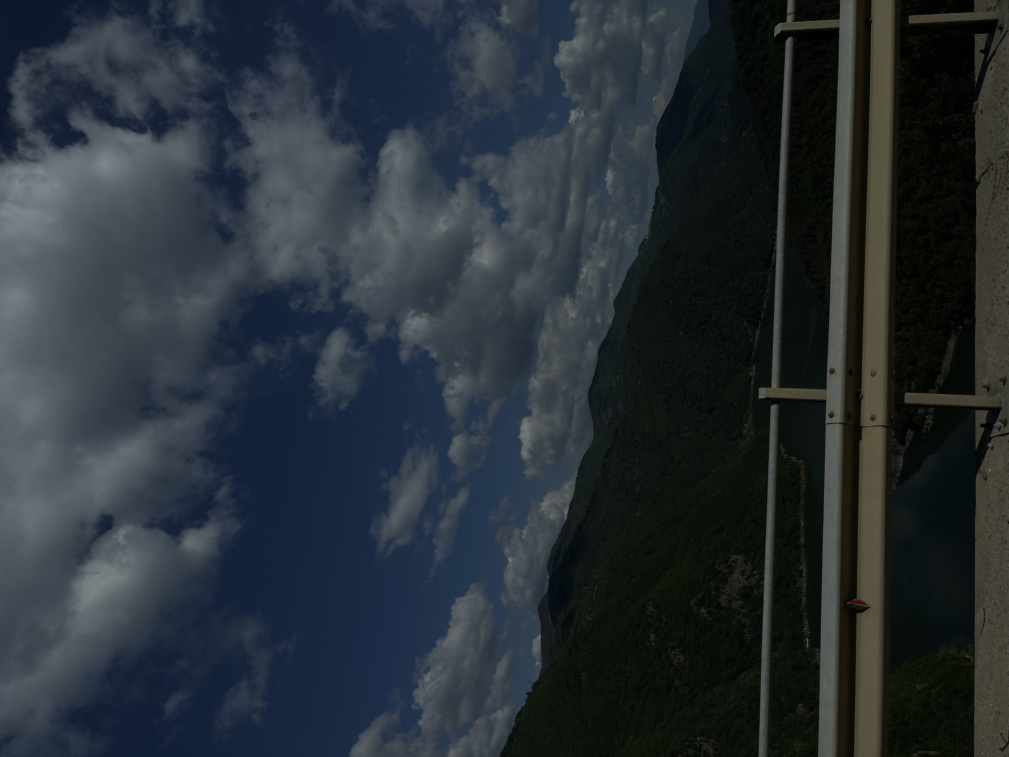 A landscape photo shows mountains and clouds from an elevated point. A light-colored metal railing stands on the right side. The mountain slopes are covered in lush, green vegetation. To the left is a vast sky filled with dark, puffy clouds. A river sits between the mountains.