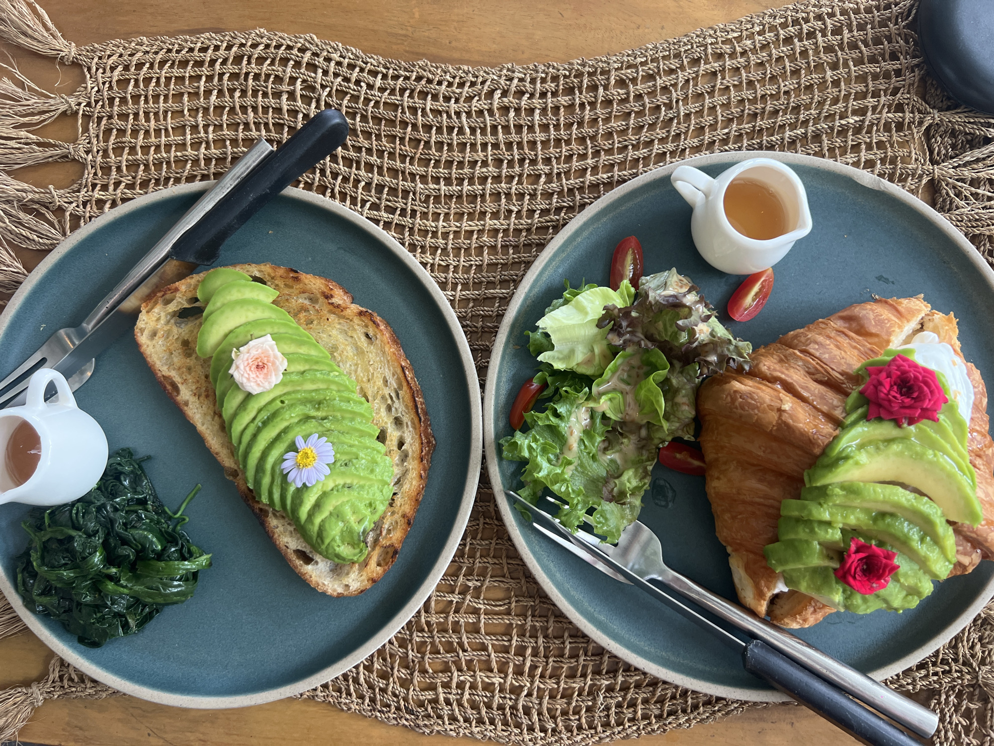 Two teal plates are set on a woven placemat on a wooden table. Each plate contains a breakfast meal with avocado. The plate on the left features a slice of toasted sourdough bread topped with fanned avocado slices and a small pink flower. To the left of the toast is a small white pitcher filled with a golden liquid, likely honey or syrup, and a serving of wilted green vegetables. A fork and knife are placed on the left side of this plate. The plate on the right holds a croissant topped with avocado slices and two small red flowers, alongside a salad of mixed greens and halved cherry tomatoes. A small white pitcher filled with golden liquid is to the top of this plate. A fork and knife are positioned on the right side of this plate. The scene suggests a breakfast or brunch setting, likely in a tropical location given the presumed setting of Baan Bo Phut, Thailand. The lighting indicates it is daytime. There is no visible text in the image.