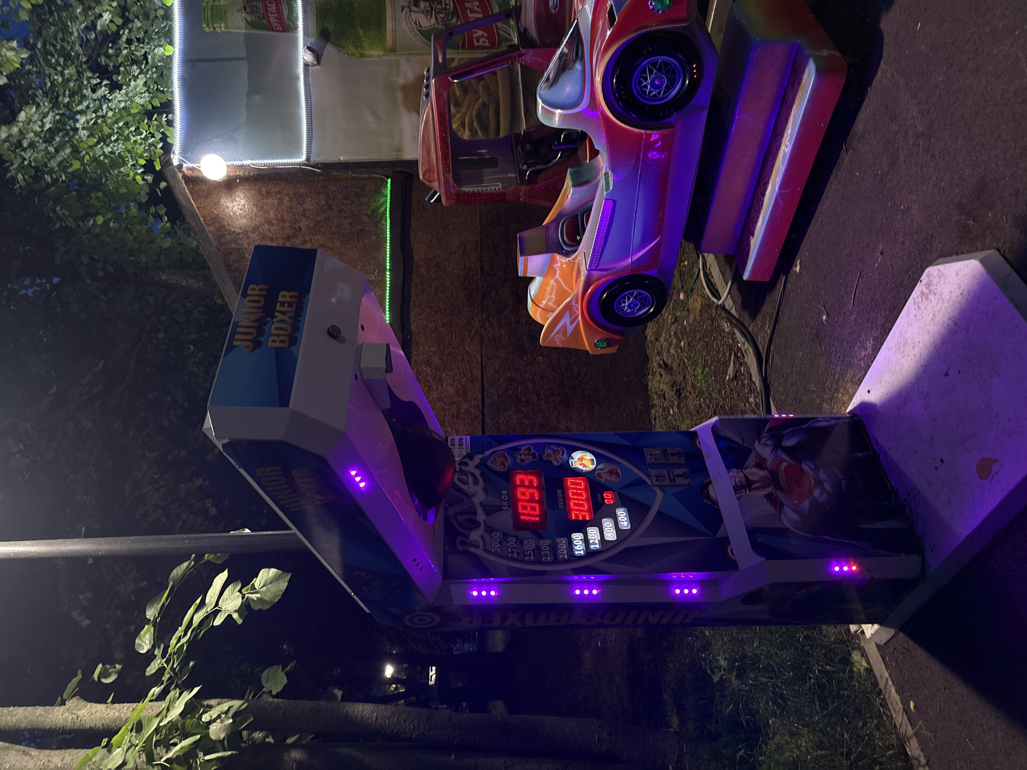 The image captures an outdoor amusement area at night, illuminated by various artificial light sources. The primary subject is a "JUNIOR BOXER" arcade punching machine, seen from an elevated perspective and angled to the right. The machine is actively lit with vibrant purple LED lights along its edges and display. Its digital readout shows a "SKOR" (score) of "1893" and a "REKOR" (record) of "3000", with other scores listed for reference. Text on the machine also includes "JUNIOR BOXER".
To the right of the punching machine, two coin-operated children's ride-on cars are visible. One is a red car and the other is an orange and red sports car, both featuring illuminated wheels. These rides are placed on dark pavement or asphalt.
In the background, behind the arcade machines, there is a structure or wall lit by white string lights and a single bright bulb. A green string of lights is also visible near the ground. Lush green foliage and tree branches are prominent on the left and bottom portions of the image, indicating a park or garden setting surrounding the amusement area. A banner or sign in the upper background clearly displays the text "БУРГАС" (Burgas), confirming the location as Burgas, Bulgaria. Another partial text "3 ИГРИ" (3 Games) is visible near the top left of the punching machine's display. The overall ambiance is lively and recreational, typical of a nighttime entertainment venue.