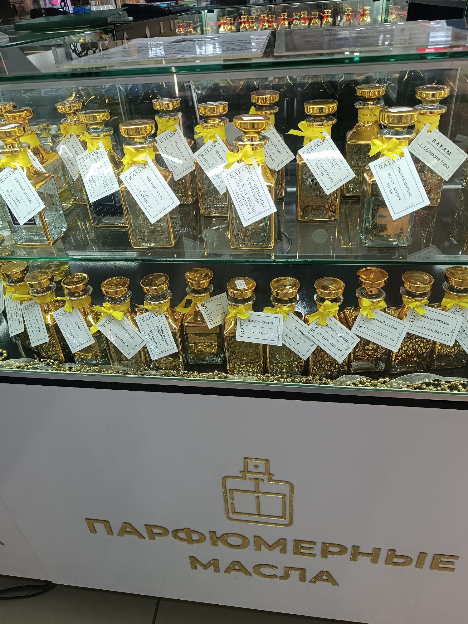 A display case containing perfume bottles labeled "RAFAM" is visible in a retail setting. The bottles are primarily square-shaped, with gold-colored accents and yellow ribbon details, each accompanied by a descriptive tag in Cyrillic and English. The bottles are arranged in two tiers. The bottom part of the case has the text "ПАРФЮМЕРНЫЕ МАСЛА" in gold letters. It is likely located in Сепыч, Russia as some of the product descriptions are in the Russian language.