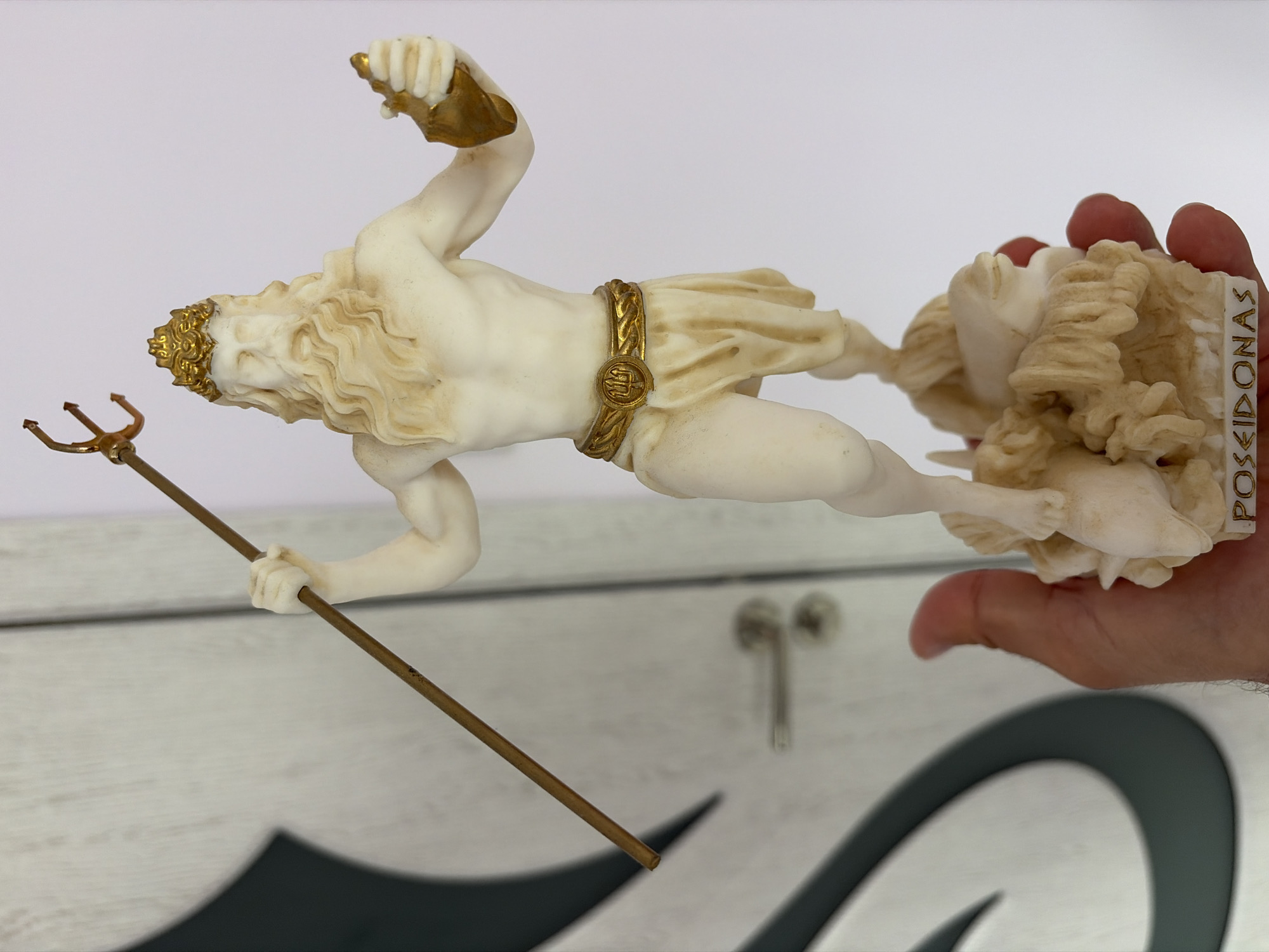 A detailed, eye-level shot captures a white and gold statuette of Poseidon held by a light-skinned hand, set against a blurred background featuring a light-colored wooden door with a dark, abstract, curved design. The statuette depicts a muscular, bearded male figure with long, wavy hair, wearing a golden crown and a golden belt adorned with a trident symbol. He holds a slender, golden trident in his raised right hand. His left hand holds a golden, cup-like object. The lower part of the statuette shows sculpted waves, marine life, and the inscribed word "POSEIDONAS" on the base. The hand holding the statuette is visible from the palm side, with fingers partially obscuring the base. The background door also shows a metallic door handle in the middle-right area.