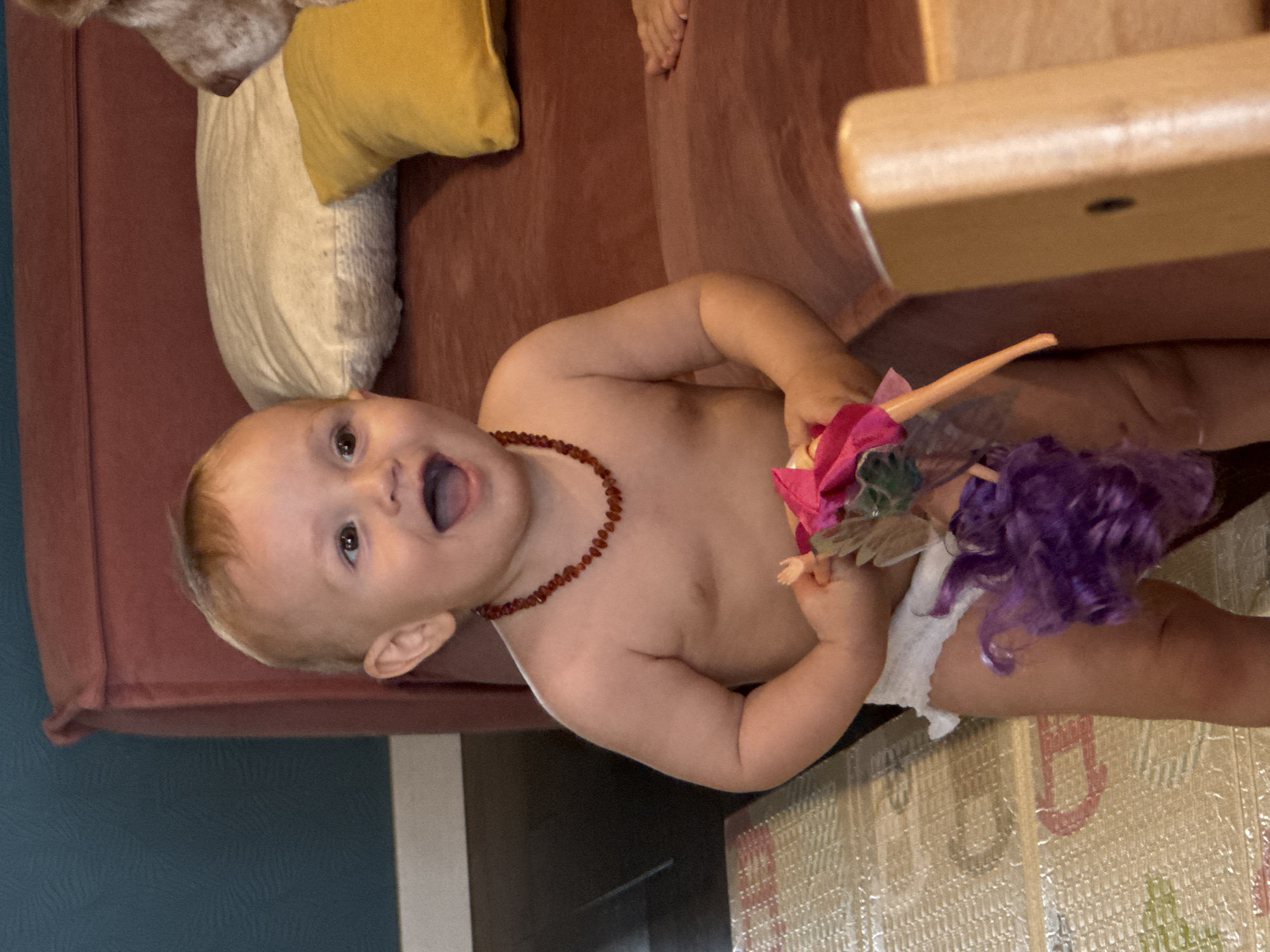 A baby wearing a diaper and a beaded necklace is holding two dolls. The baby is standing on a corrugated mat near a red couch with pillows. A part of a wooden chair is visible. The location is unknown and cannot be determined to be in Moscow, Russia.