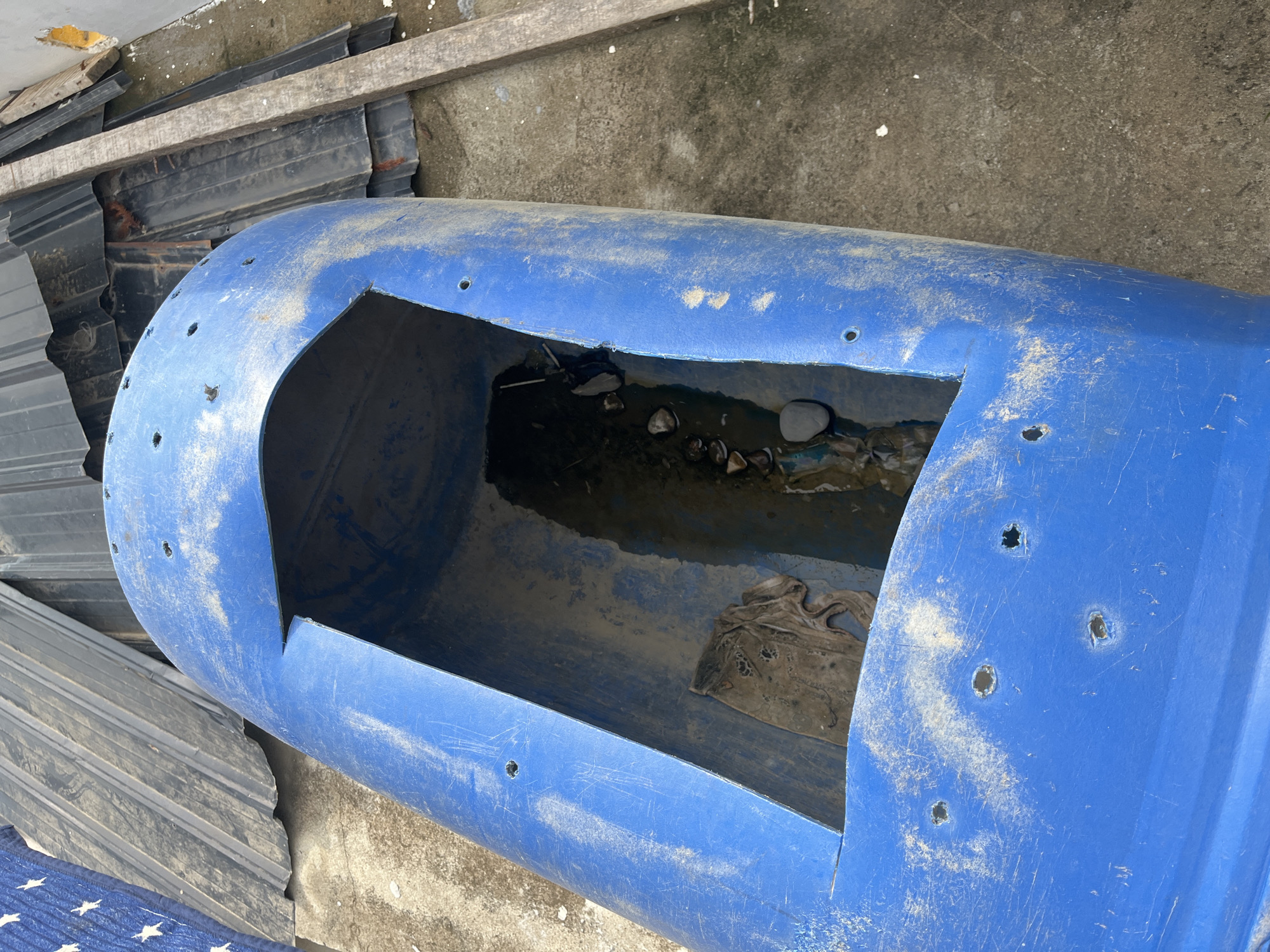 In Benuaraja, Indonesia, a blue, cylindrical plastic container is situated on a concrete surface. The container is approximately two-thirds submerged in murky water, with a piece of dirty, light-colored fabric visible inside. The container has a large rectangular opening cut into its top, revealing the water and debris within. Numerous small holes are drilled into the outer surface of the container. To the left and behind the container, corrugated metal sheets are stacked, some appearing rusted. A wooden plank is positioned diagonally to the right of the container. The ground is concrete, with some scattered white debris. The scene appears to be outdoors in a storage or construction area.