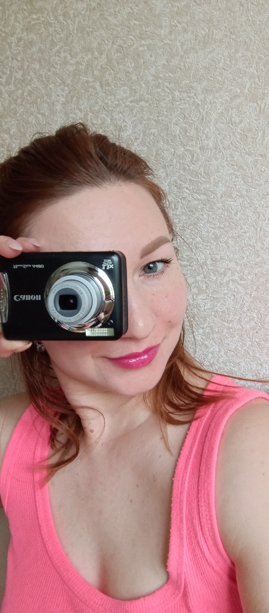A woman holds a black Canon camera up to her face, partially obscuring it. She has fair skin, blue eyes, and reddish-brown hair. She is wearing a bright pink ribbed tank top. The background is a textured, neutral-colored wall. The location is Unknown, Unknown.