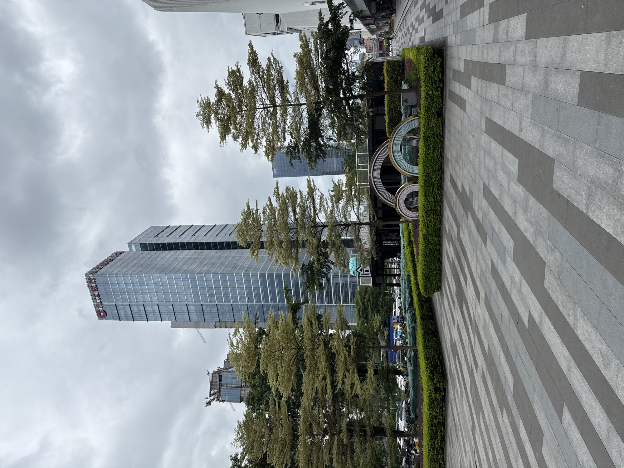 The image depicts an outdoor urban scene in Tianhe District, China, under an overcast sky. In the foreground, a wide pedestrian walkway is paved with rectangular gray and dark gray tiles laid in an alternating pattern. To the left, a manicured garden with low green bushes borders the walkway. Behind the garden, several trees with sparse, light green foliage are planted.
Dominating the background are modern high-rise buildings. The most prominent is a tall, slender glass skyscraper with a red logo at its apex. To its left, another skyscraper under construction is visible, featuring a crane and scaffolding. Further to the right, another glass building with a more curved facade stands.
In the mid-ground, a public art installation is visible within the landscaped area. It consists of three large, semi-circular structures, two of which appear to be made of iridescent, segmented materials, while the third is partially obscured. A low hedge surrounds the art installation and the garden area. A parking lot with several cars and motorcycles is visible behind the trees and hedges. A section of a building with some windows and a blue circular object is also present to the right of the art installation. The overall impression is of a modern, developing urban area with a mix of commercial and green spaces.