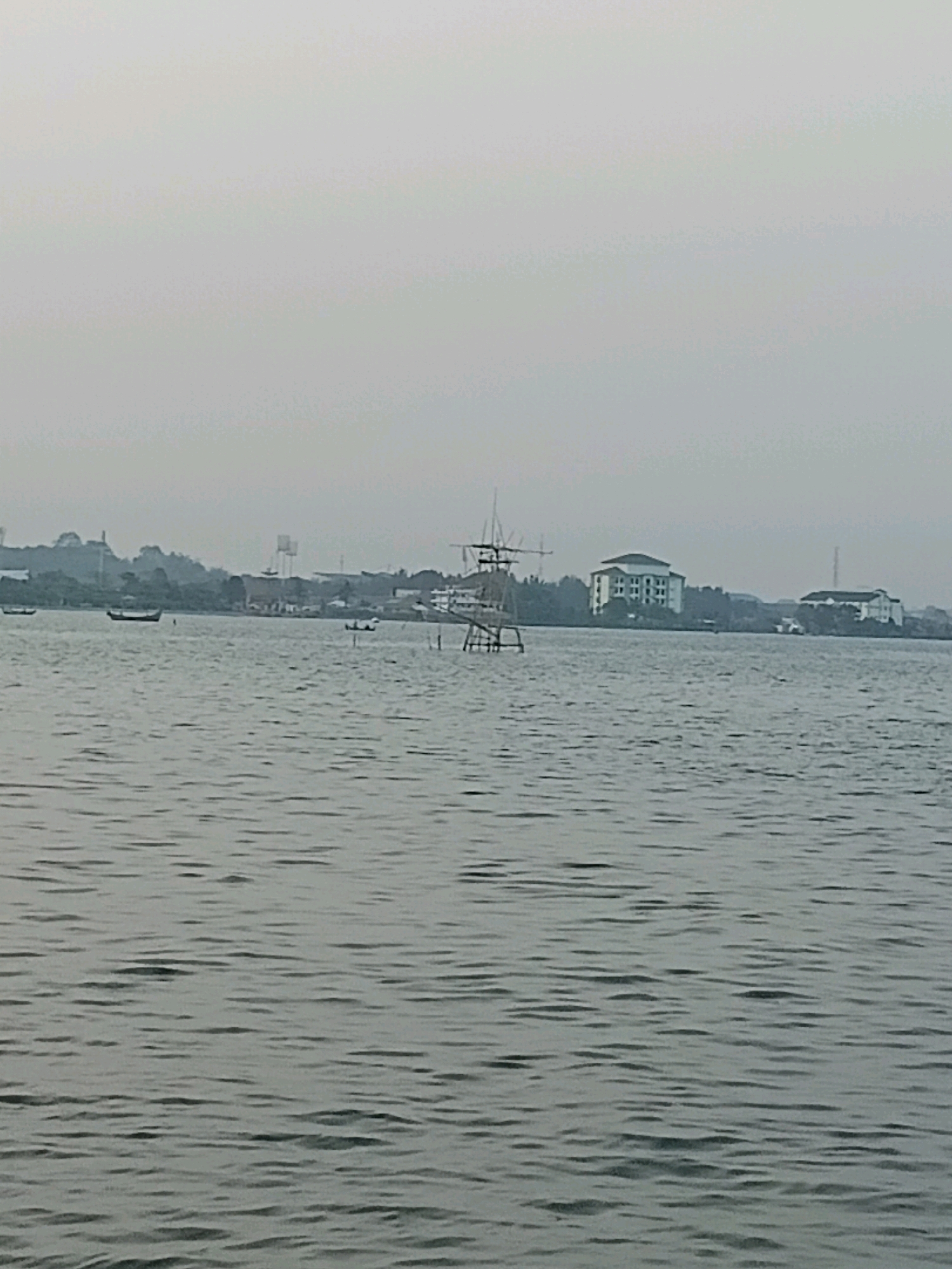 The image shows a body of water, likely a river or bay, with a hazy sky. In the background, there is a distant shoreline with several buildings, including at least two multi-story structures. There is also some vegetation visible along the shore. Several small boats are scattered on the water. In the foreground and middle ground, the water has a rippled texture. A tall, skeletal structure, possibly a fishing platform or observation tower, stands in the water on the right side of the image. The overall impression is of a coastal or riverside urban environment under overcast conditions.